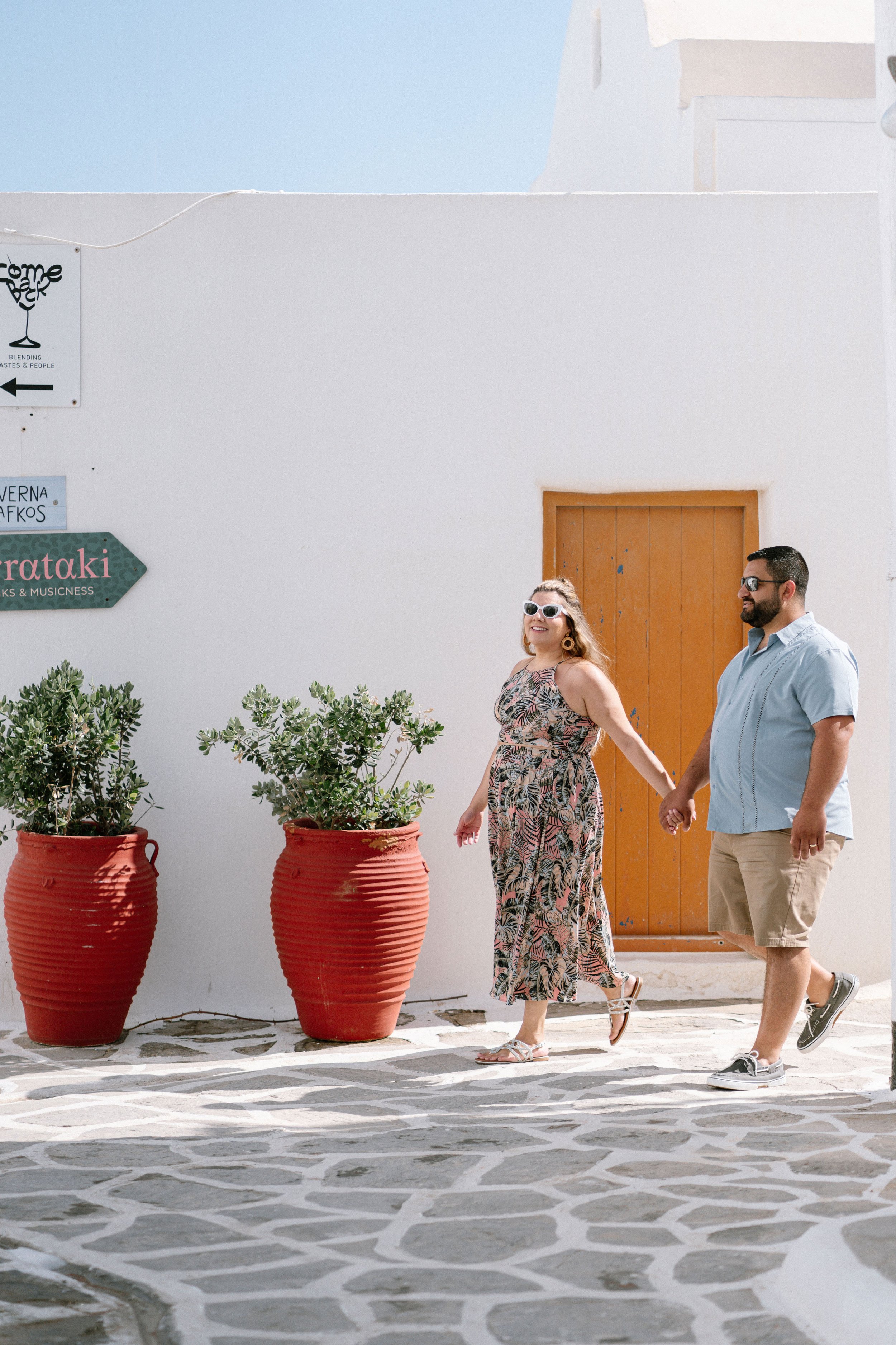 A couple holding hands walking on a sunny street with white buildings and large red planters with greenery, in a Mediterranean setting.