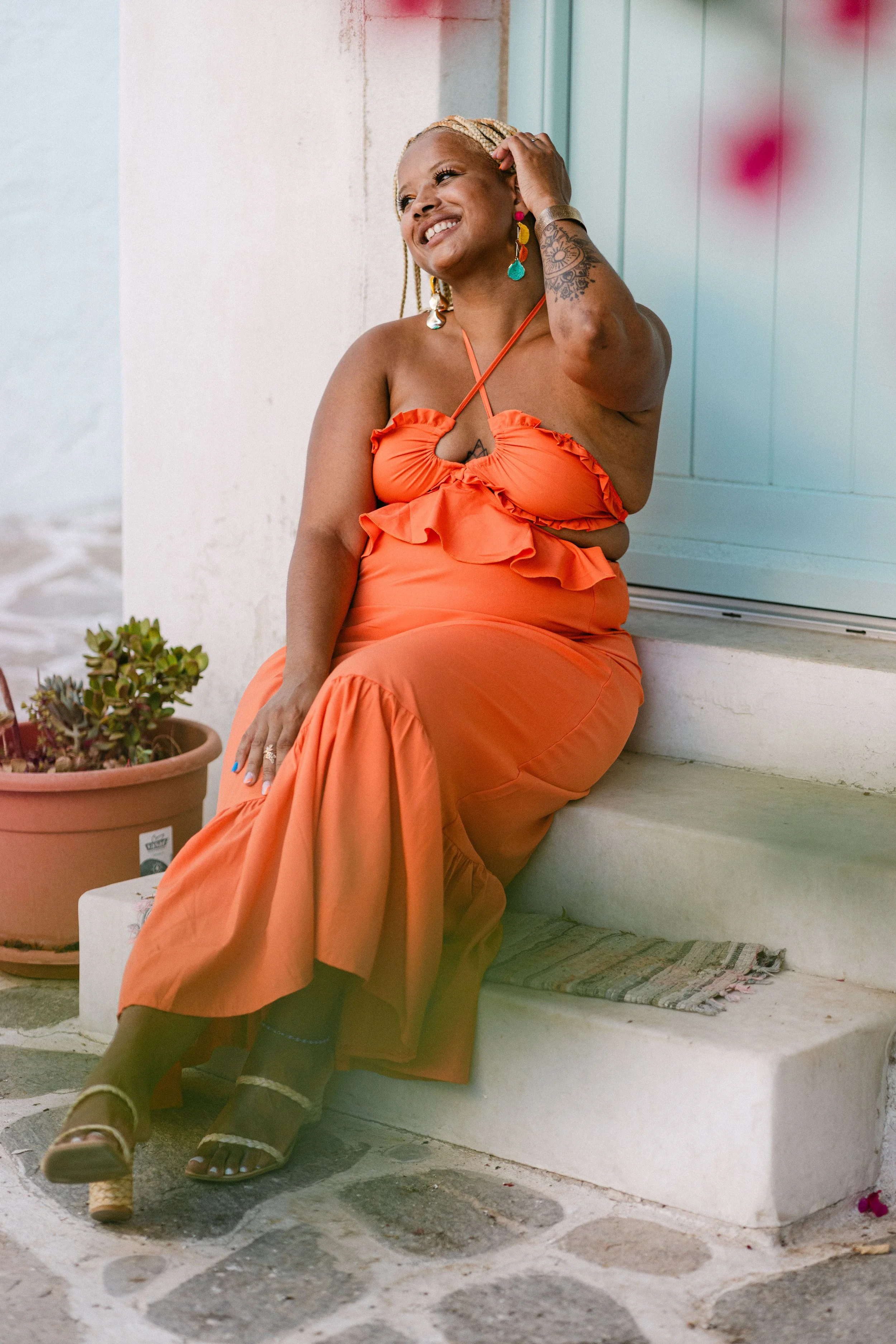 A woman sitting on a white step outside, wearing an orange dress, laughing and touching her hair, with a potted plant nearby.