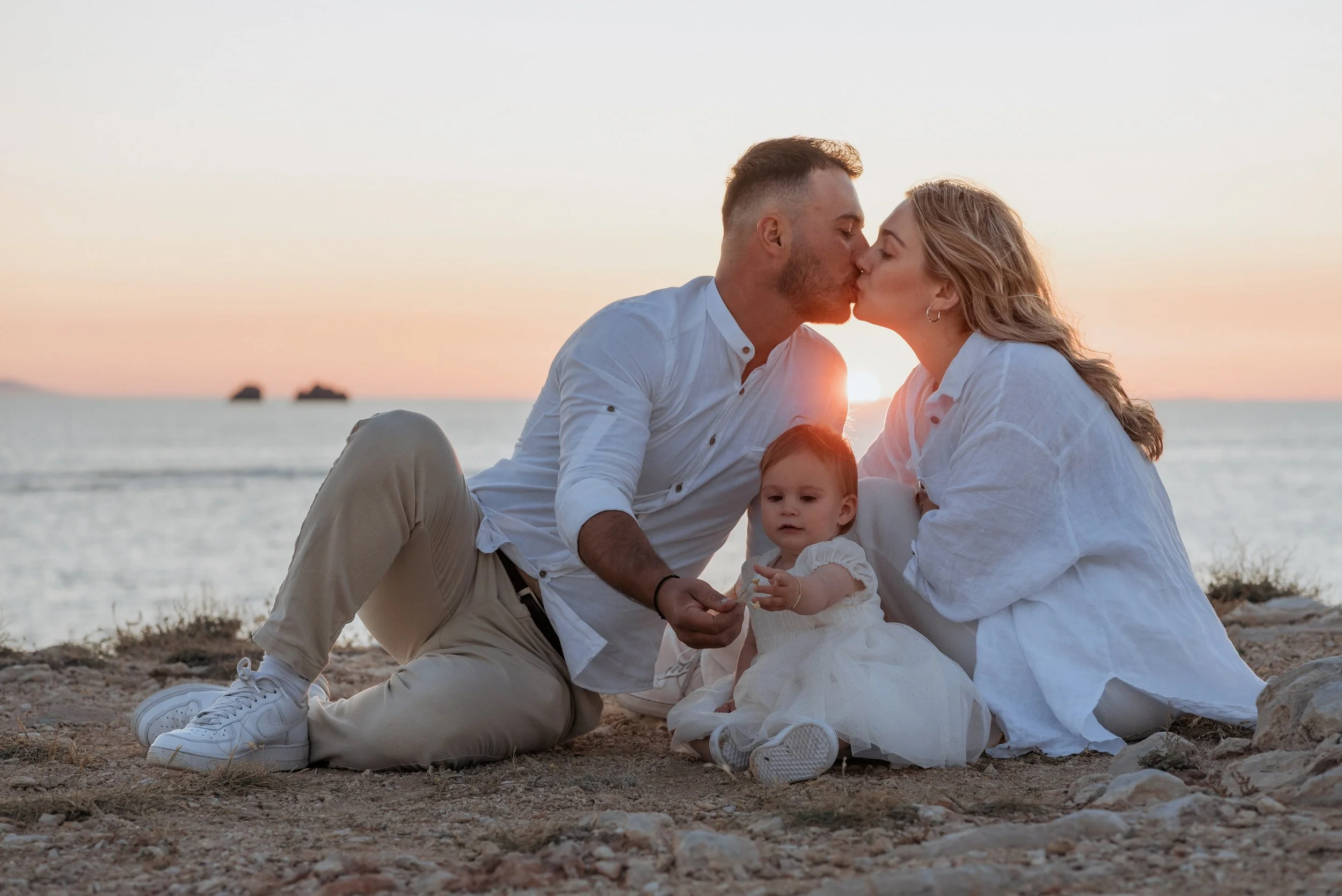 A family of three, a man, woman, and a young girl, sitting on a beach at sunset. The man and woman are kissing, and the young girl is sitting between them, holding hands with the man. The family is dressed in light-colored, casual clothing, and the b