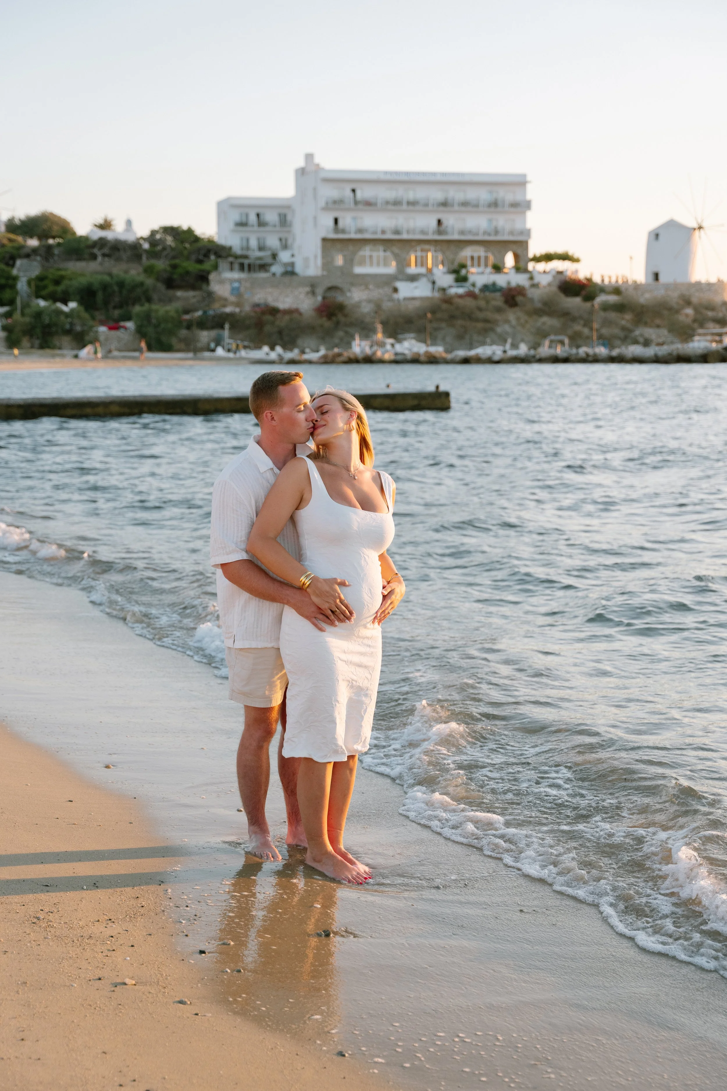 A couple standing in the shallow water at the beach, sharing a kiss, with a white building and a windmill in the background during sunset.