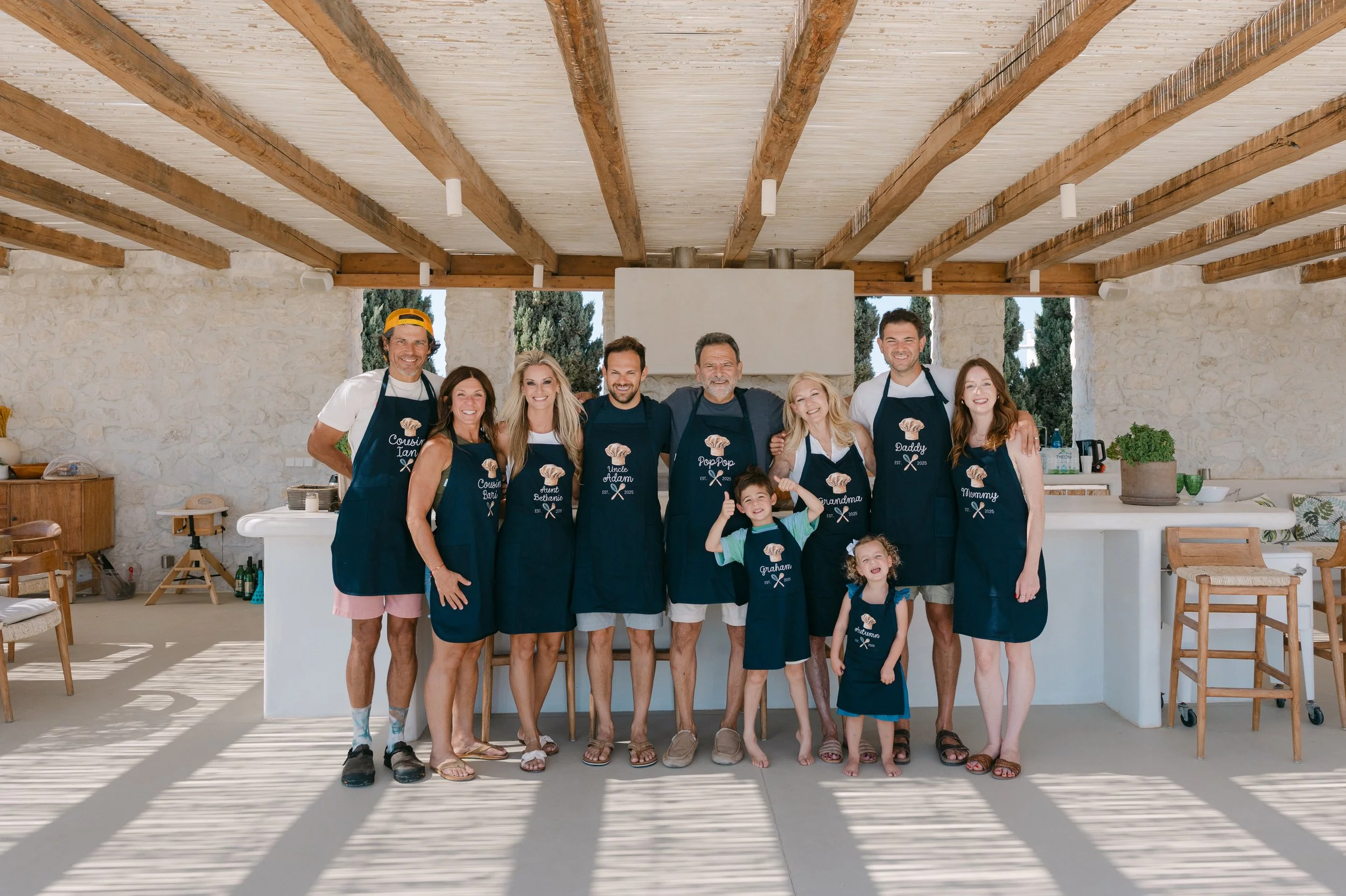 A group of eleven people, including adults and children, standing together inside a spacious, airy kitchen with a high wooden ceiling, stone walls, and large windows. They are all wearing matching navy aprons, some with names and a chef hat logo, and