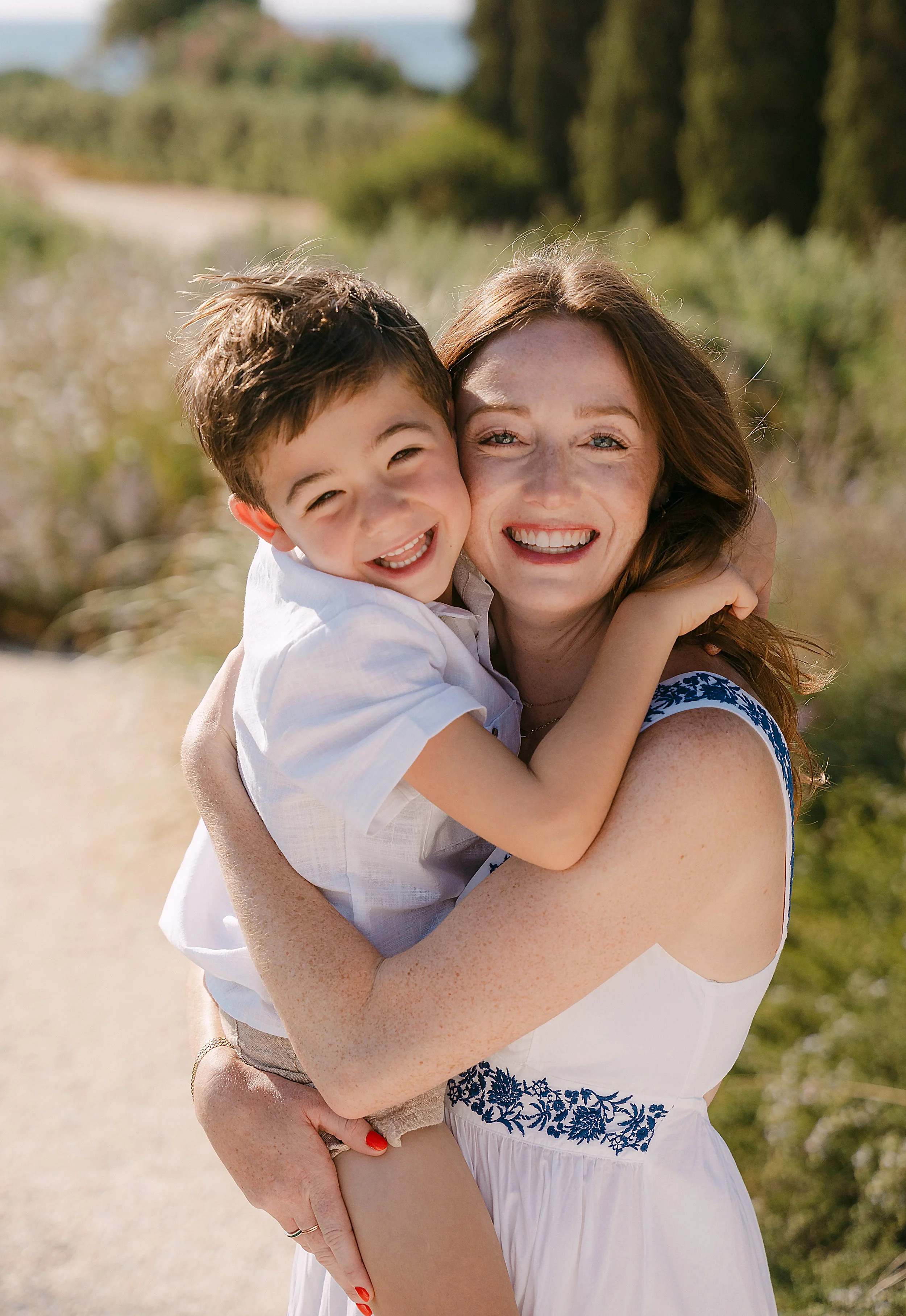 A woman with red hair and a young boy hugging and smiling outdoors on a sunny day.