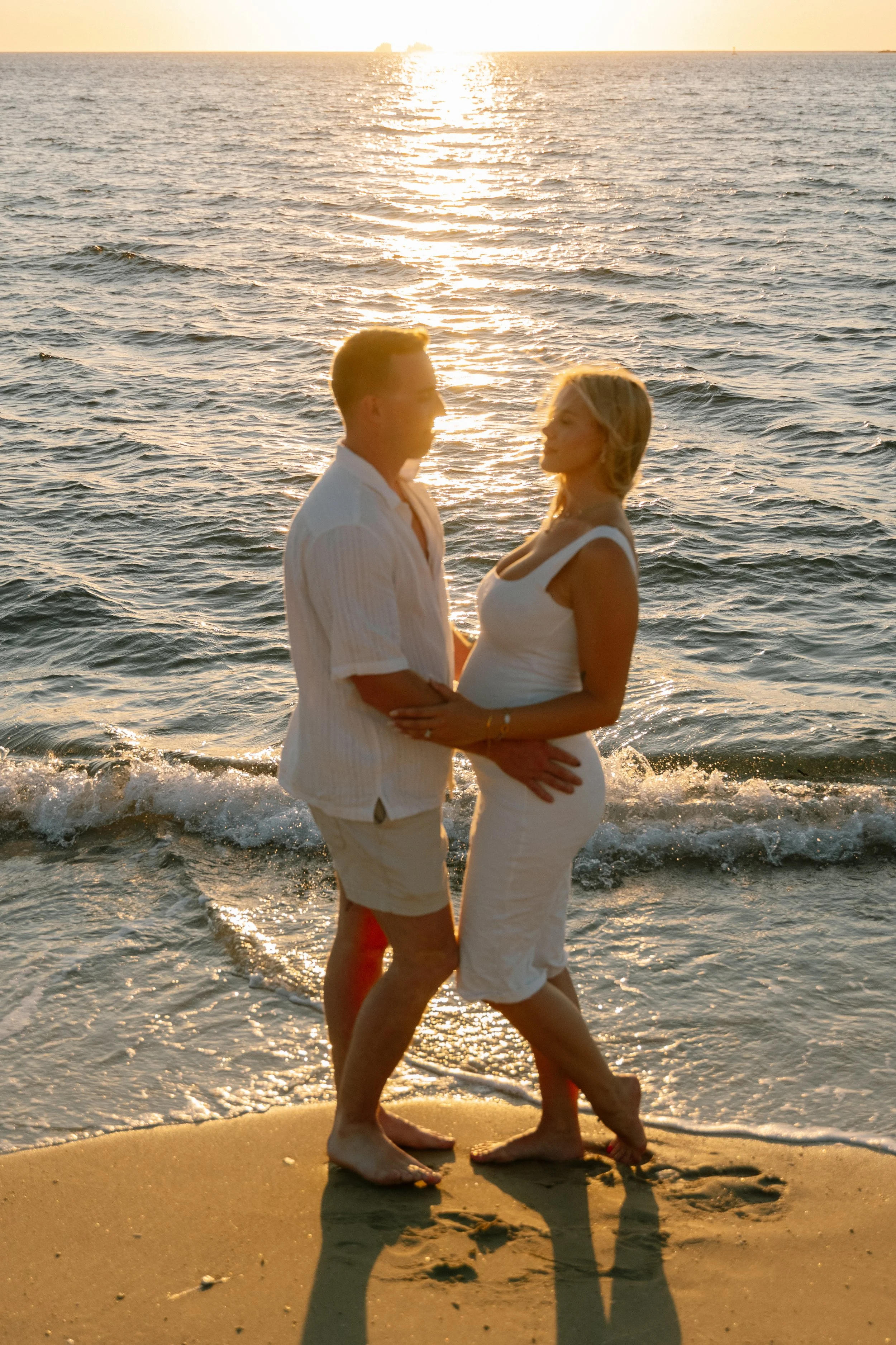 A couple standing on the beach at sunset, holding hands and facing each other.