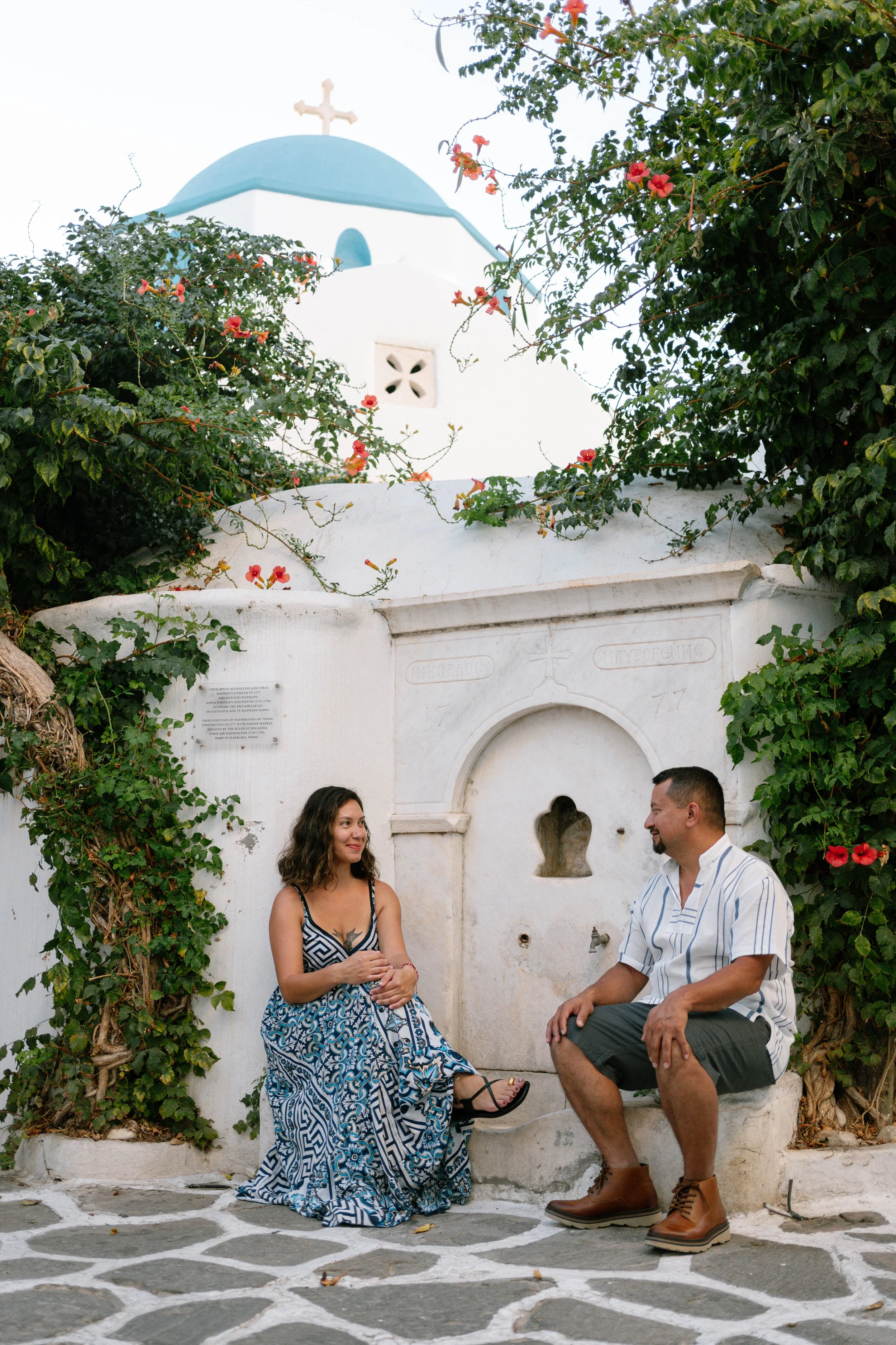 A man and woman sitting and smiling at each other in front of a white stone fountain with vines and red flowers, with a blue-domed building topped with a cross in the background.
