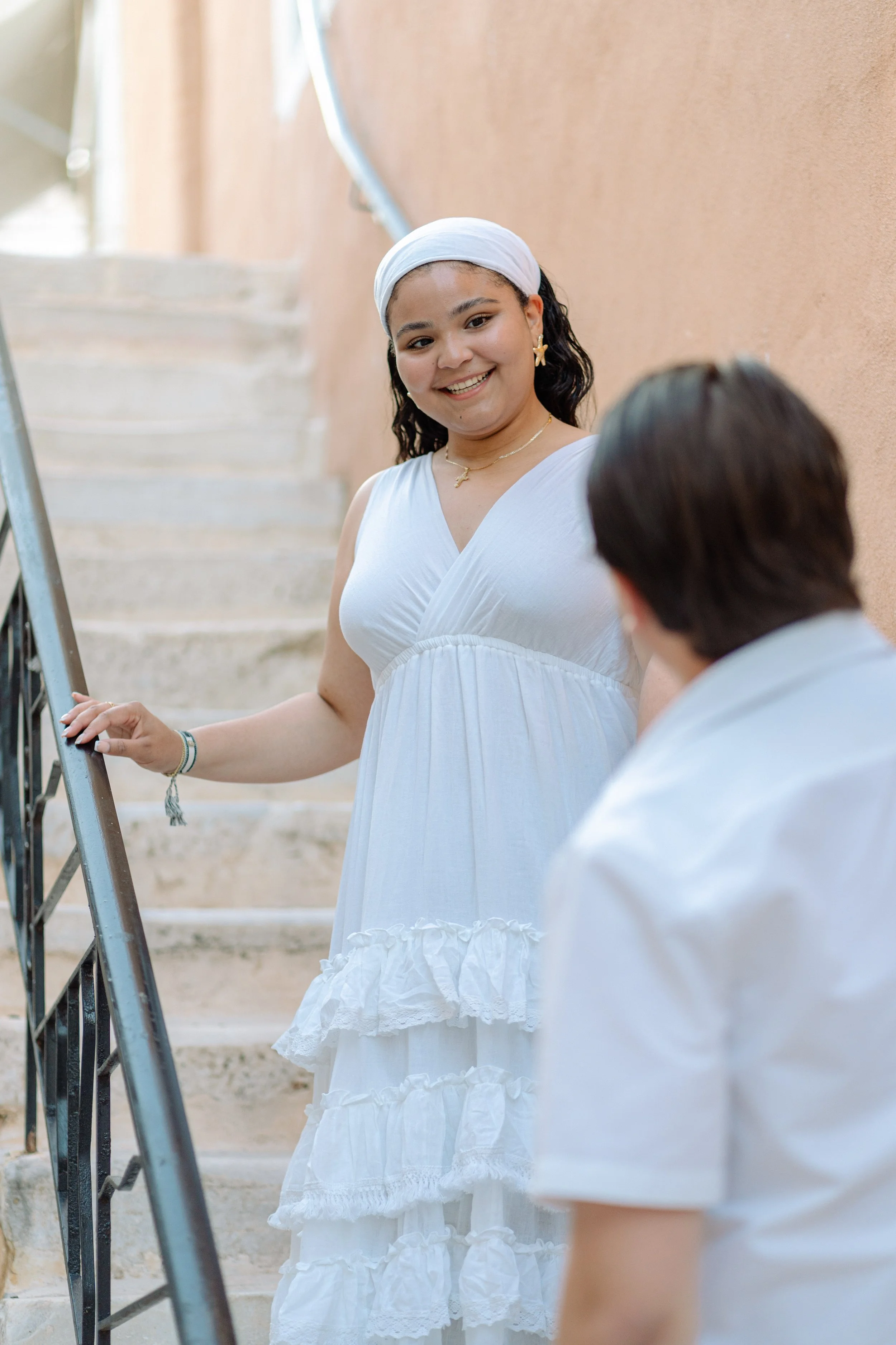 A woman in a white dress and headscarf smiling at a person with short dark hair, near a staircase and orange wall.