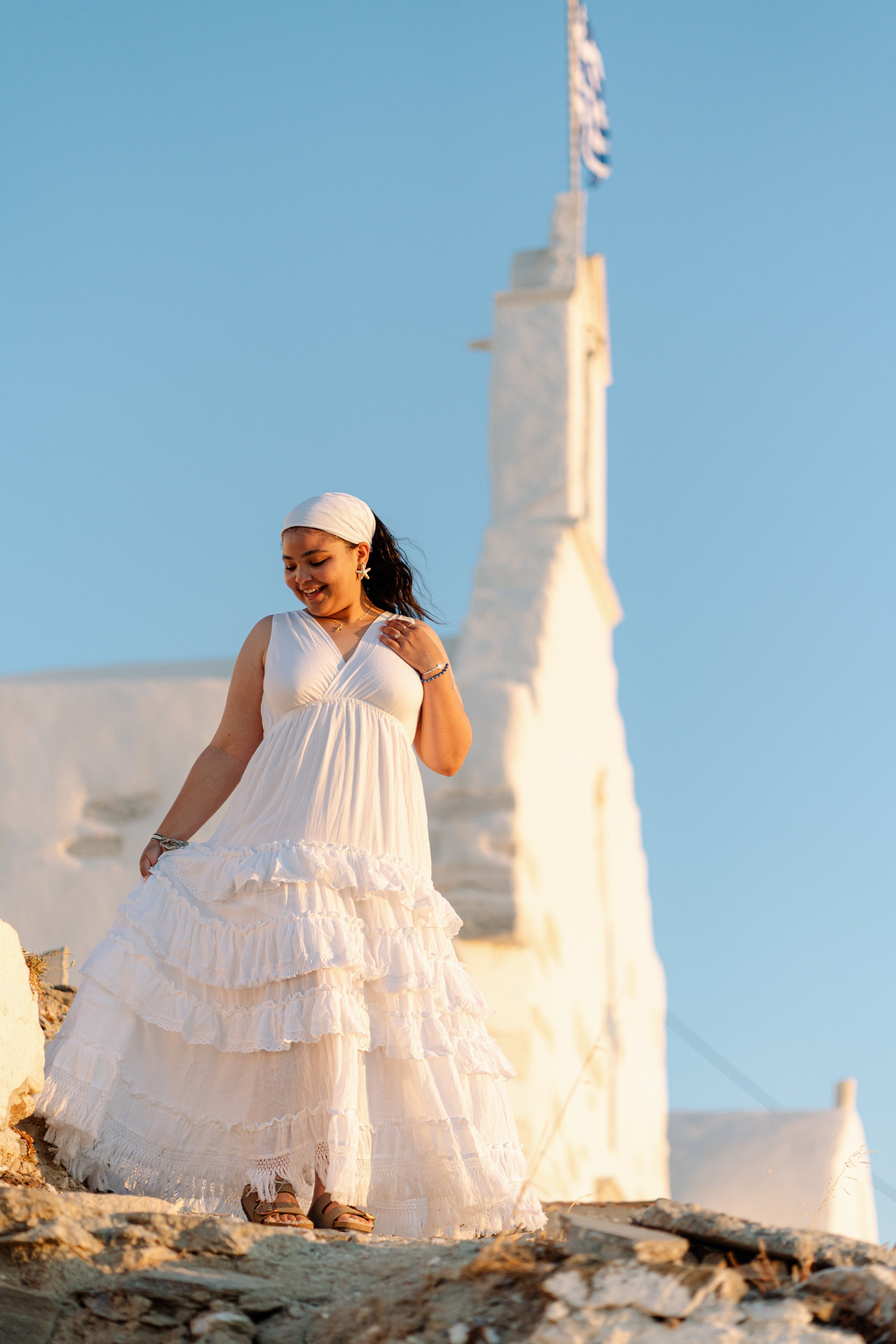 A woman in a white, flowing dress and headscarf smiling while holding her dress, standing on rocky ground with a white building and blue sky in the background.