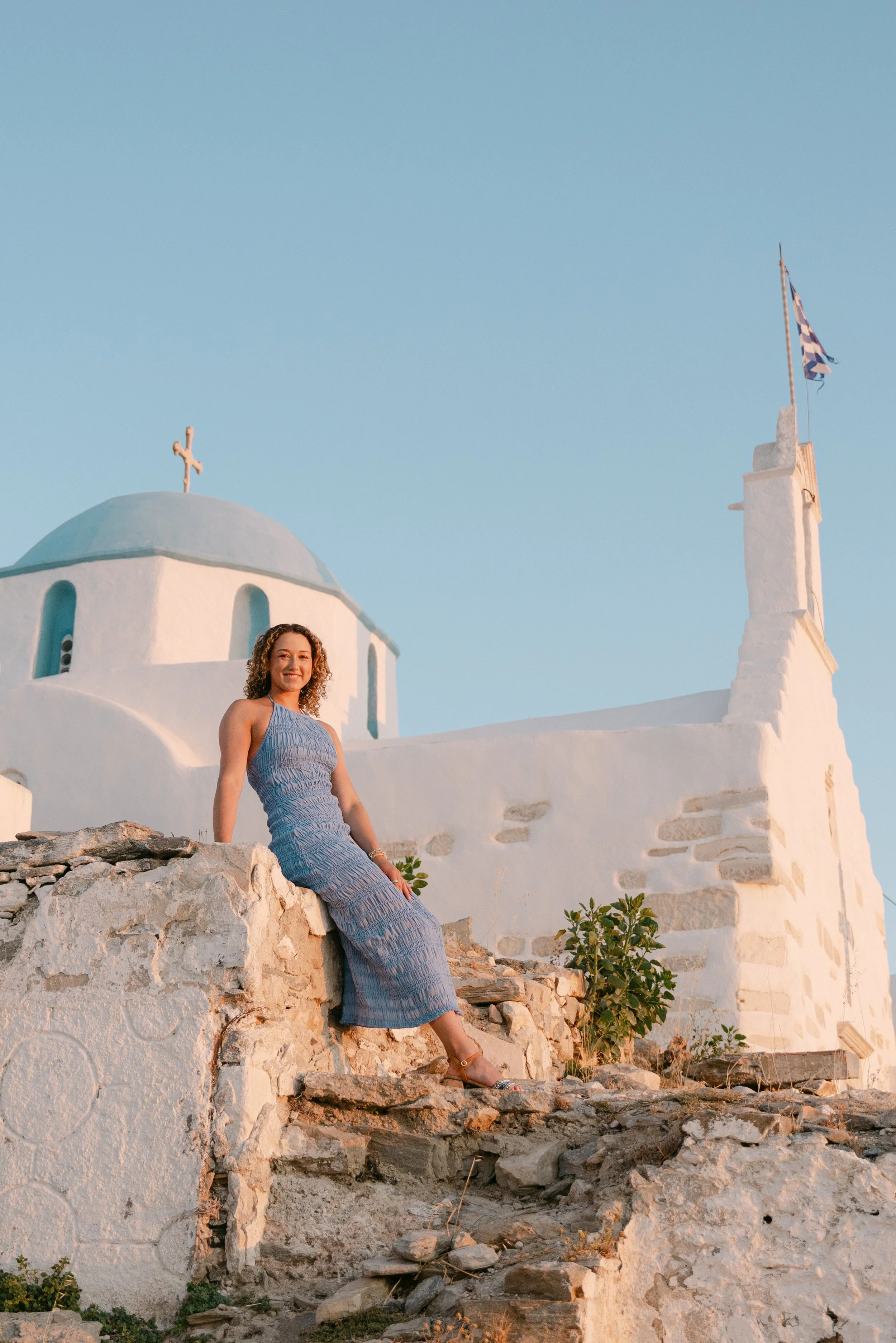 A woman in a blue dress sitting on a stone wall with a white Greek church with a domed roof and cross in the background, under a clear blue sky.