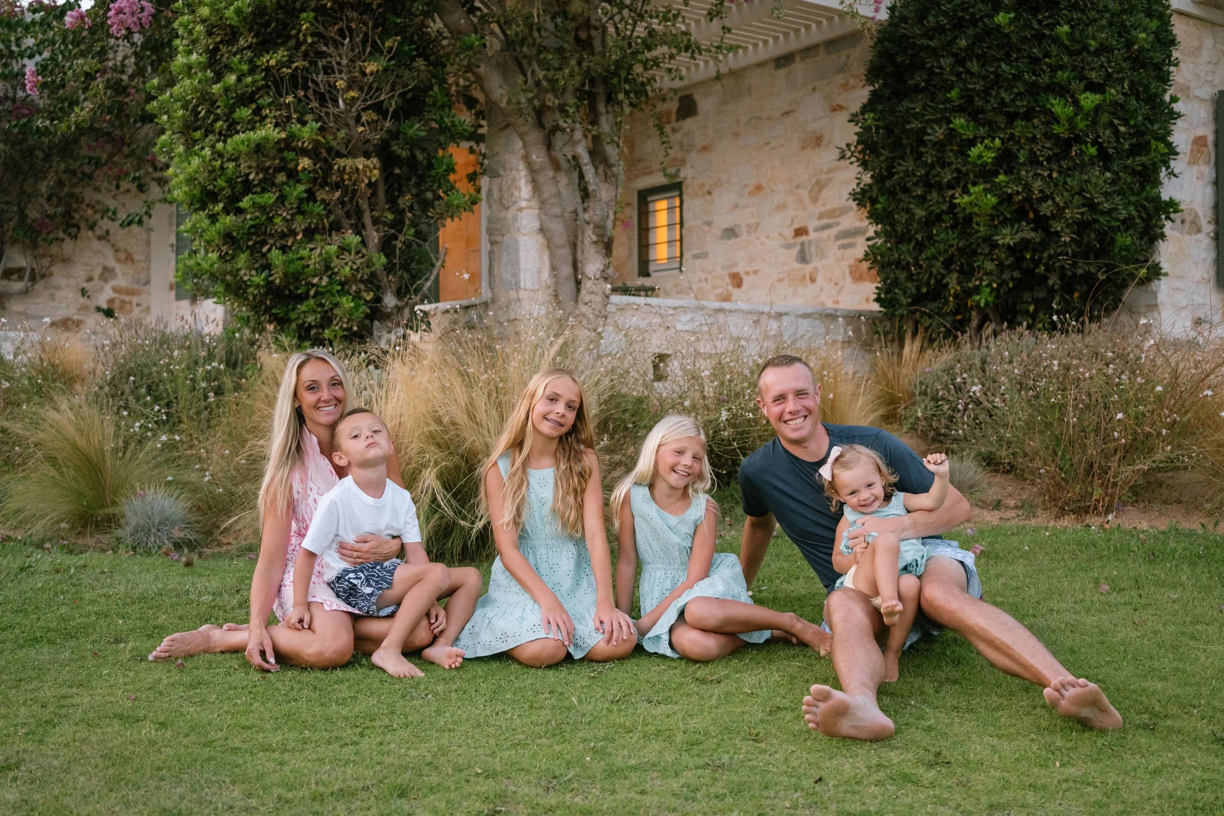 Family of six, including two adults and four children, sitting and lying on grass in a garden with plants and a stone house in the background, smiling and enjoying time together.