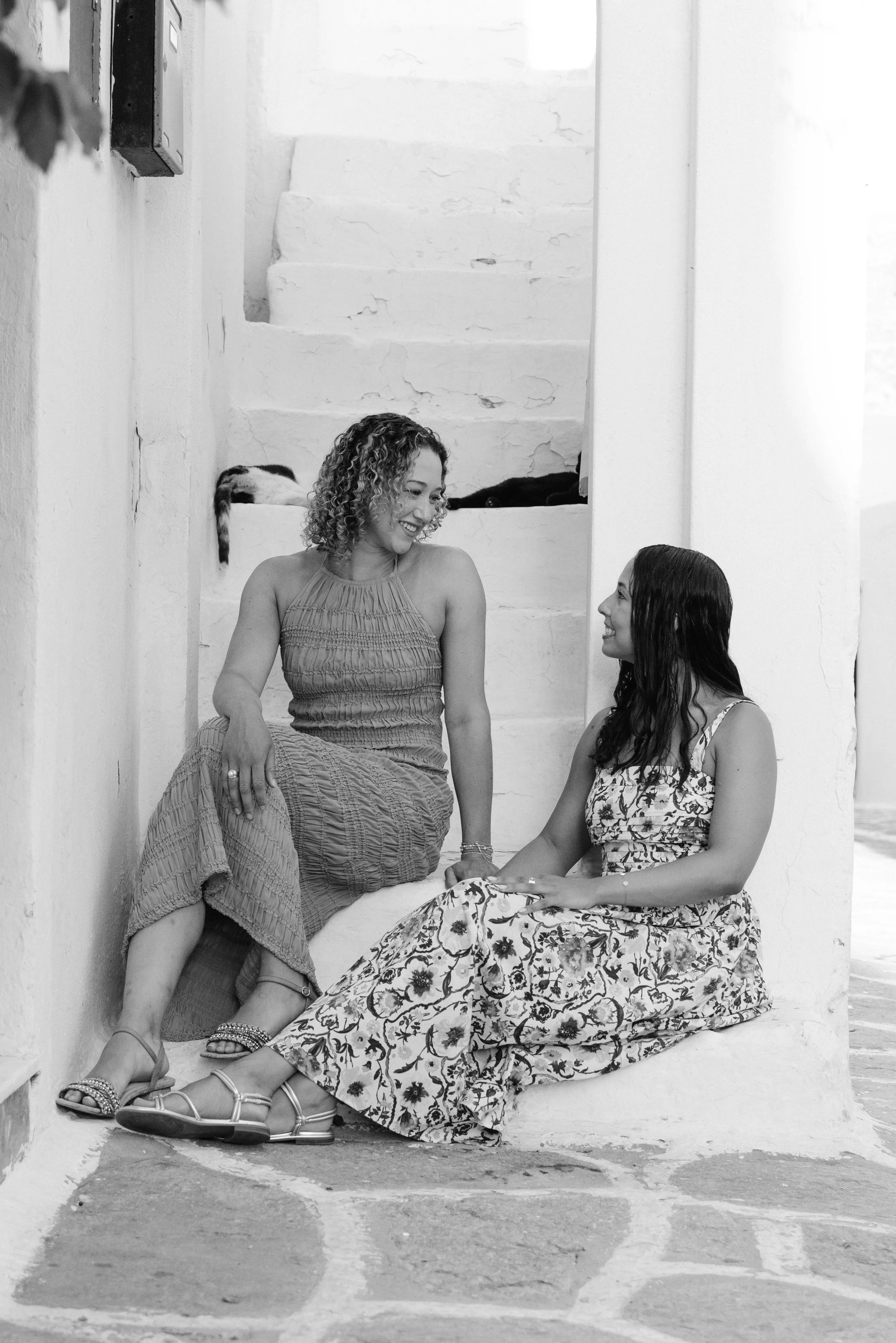 Two women sitting on a step, smiling and talking to each other, in a bright, white-washed area with a staircase behind them.