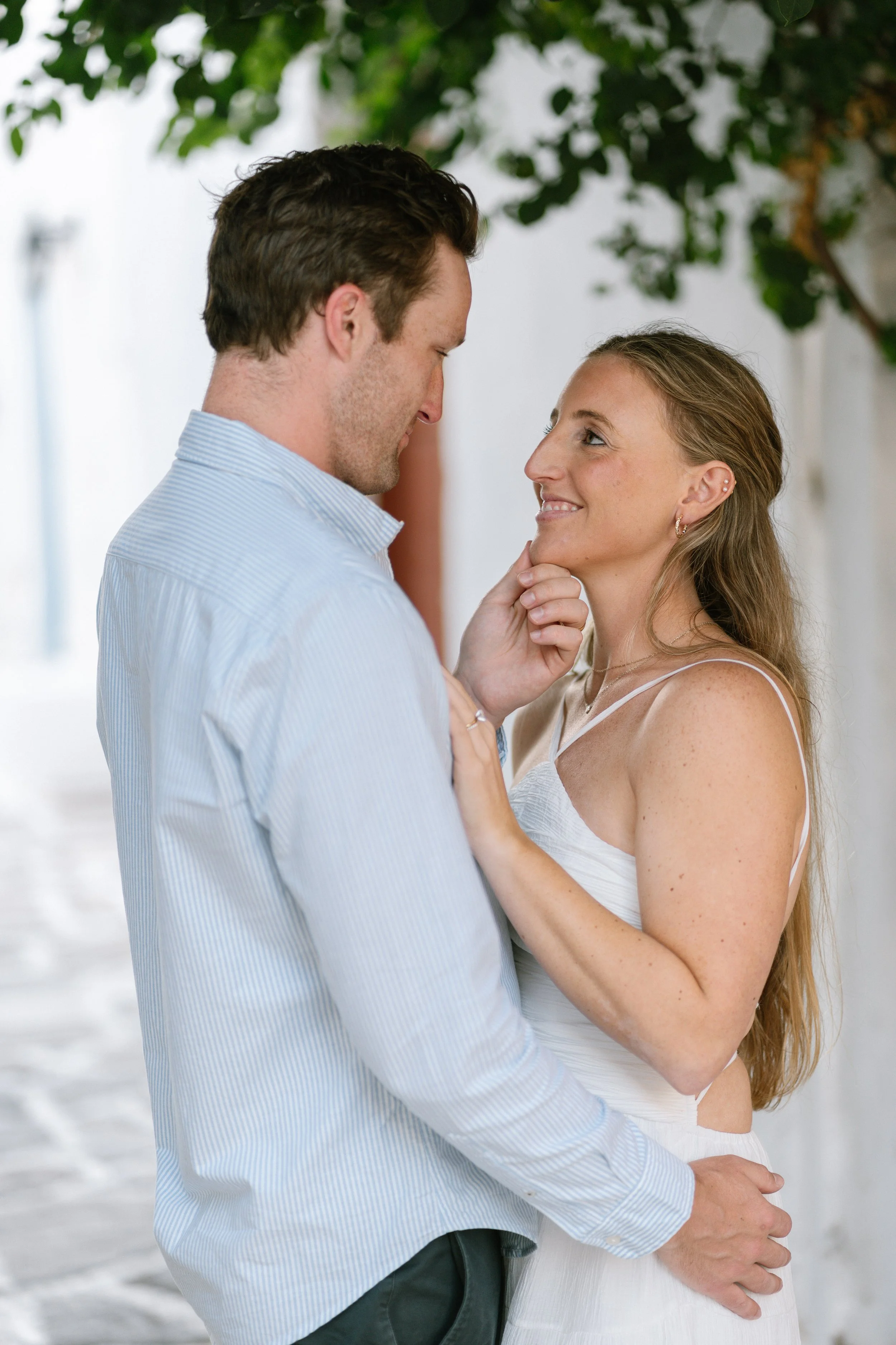 A couple gazes into each other's eyes, smiling, with the man's hand on the woman's waist and her hand on his chin, in an outdoor setting with trees and a white wall.