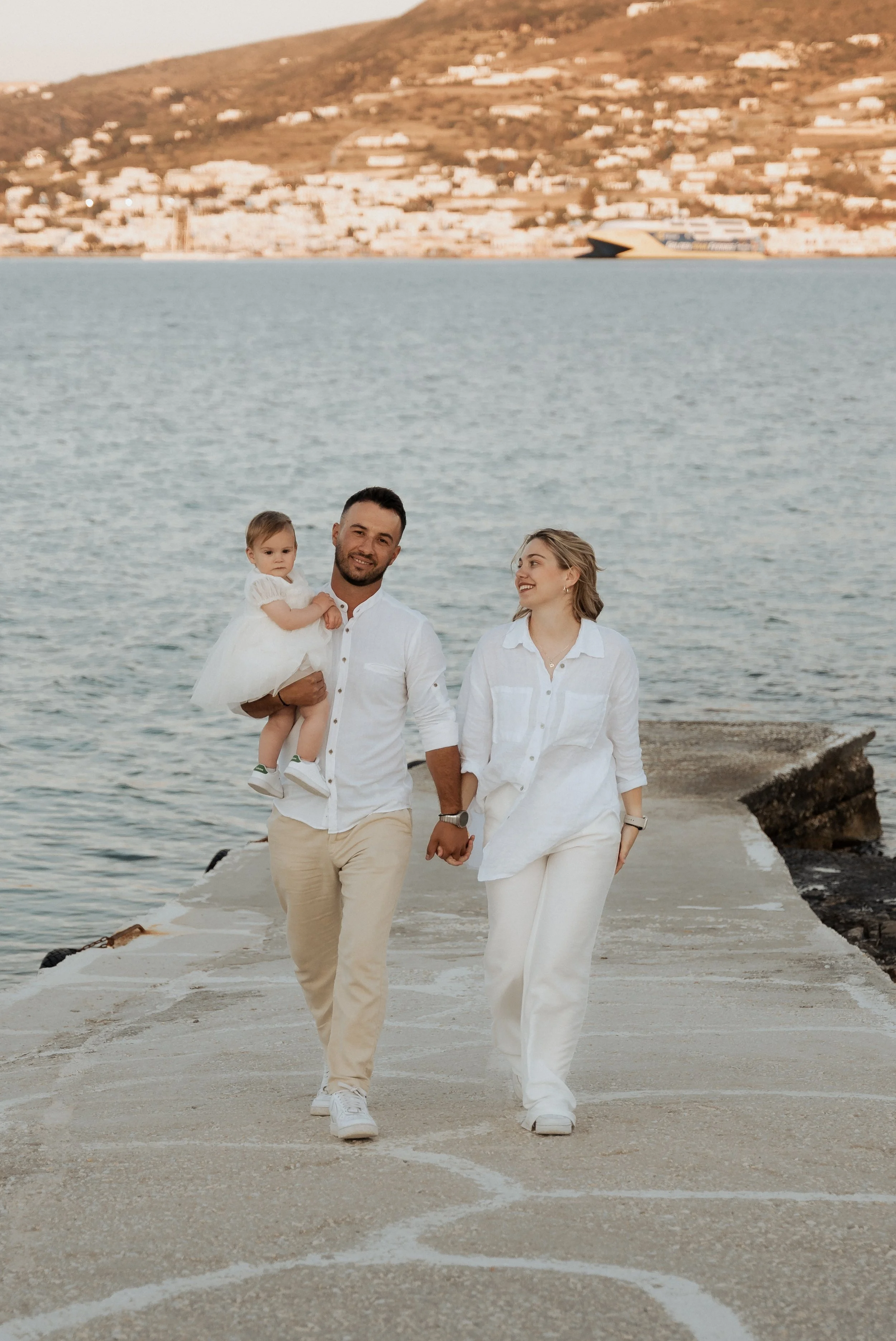 A family of three walking on a beach pier holding hands, with a man carrying a young girl in a white dress, and a woman walking beside them, all dressed in white, with water and hills visible in the background.