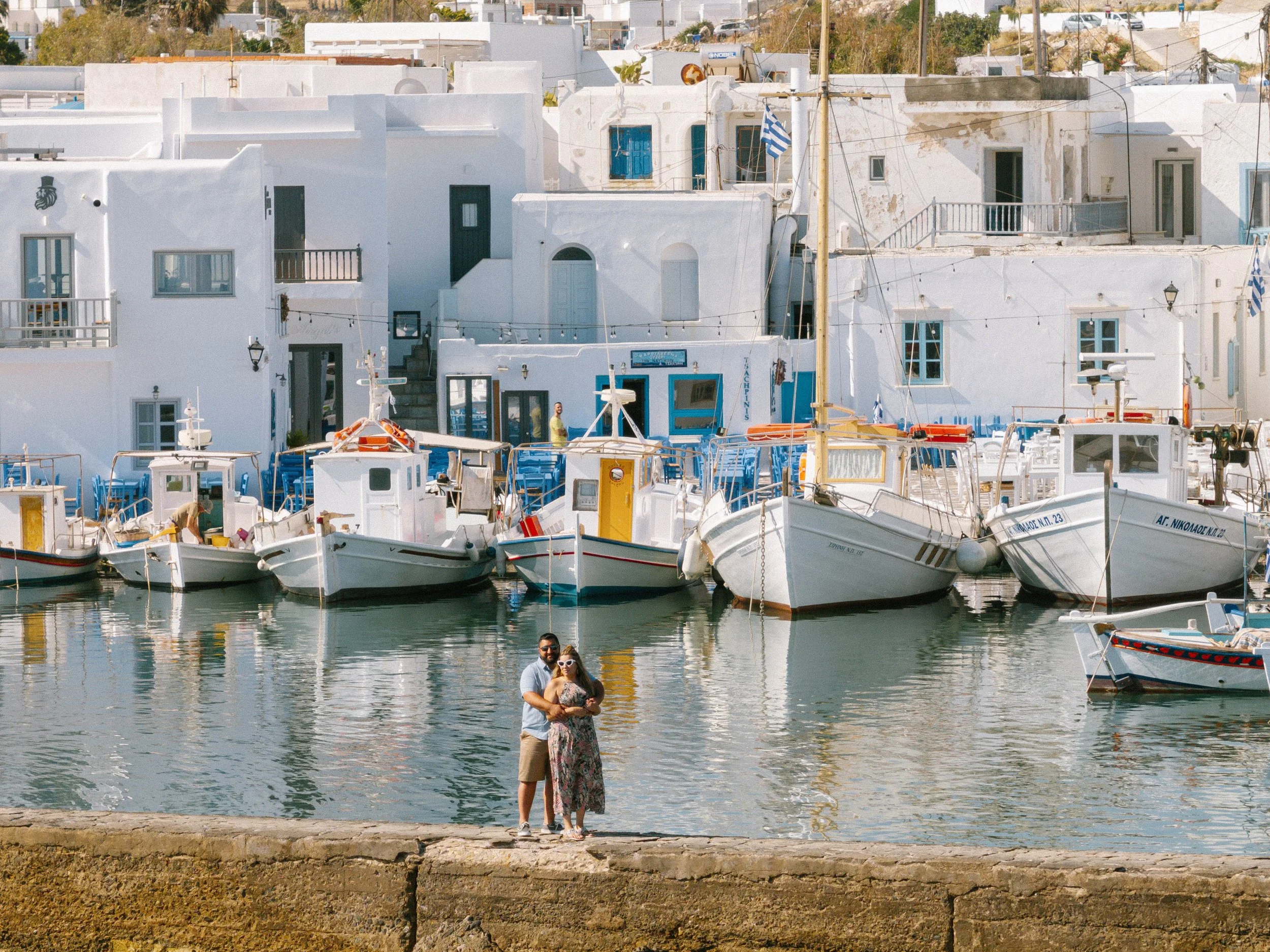 A couple standing by a waterfront with white buildings and boats docked in a harbor, sunny weather, possibly in a Mediterranean town.