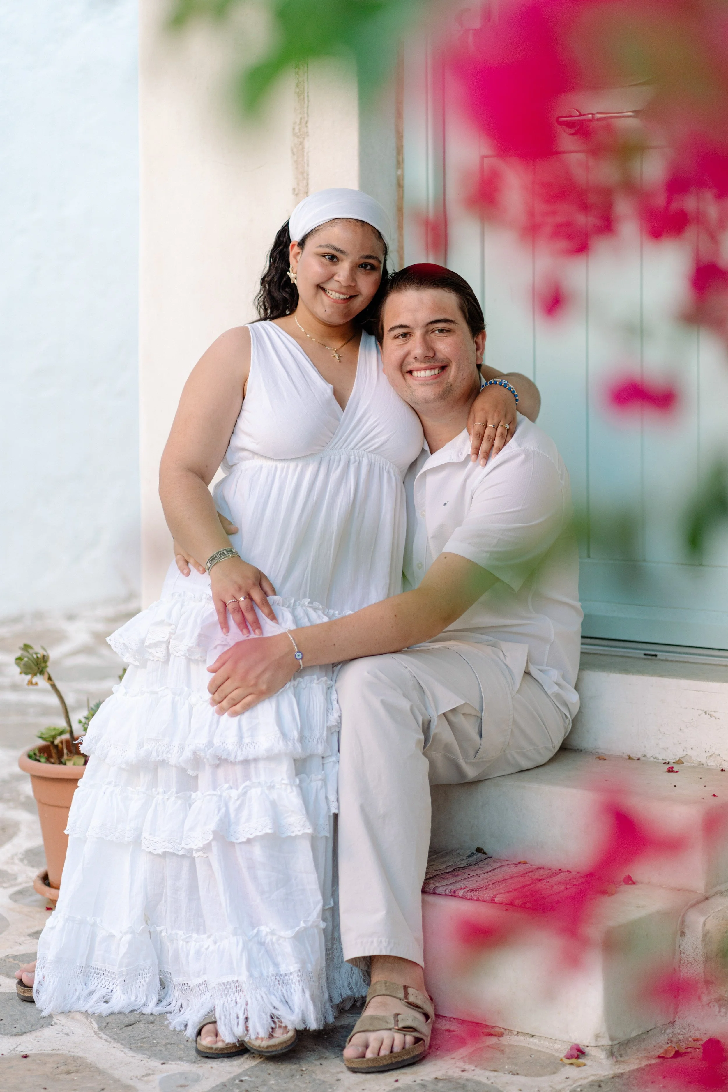 A happy couple, with the woman sitting on the man’s lap, smiles at the camera outside near a white building. The woman wears a white dress and headscarf, and the man is dressed in a white shirt and beige pants. Pink flowers are blurred in the foregro