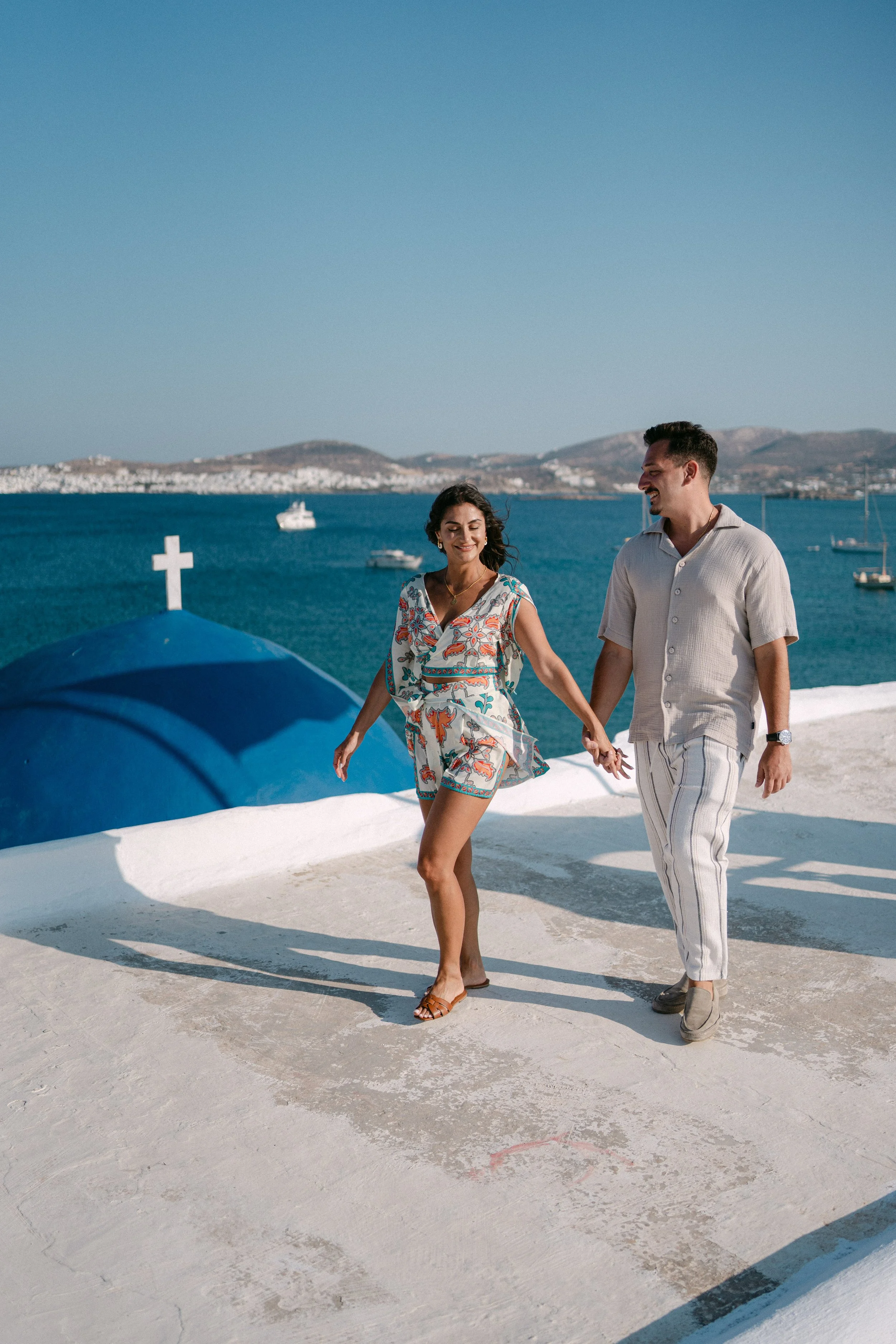 A couple holding hands and dancing on a white rooftop near a blue church with a cross, overlooking a body of water with boats and hills in the background.