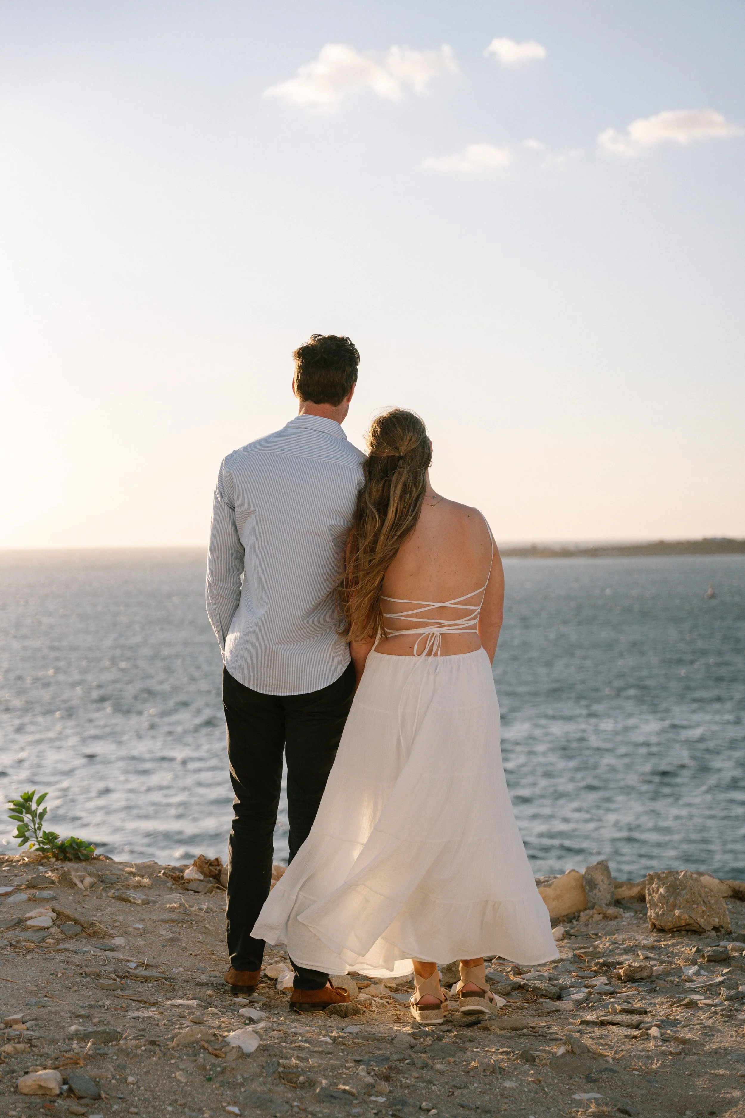 A couple stands on a rocky shore by the water, facing away from the camera, with the sky and ocean in the background during sunset.