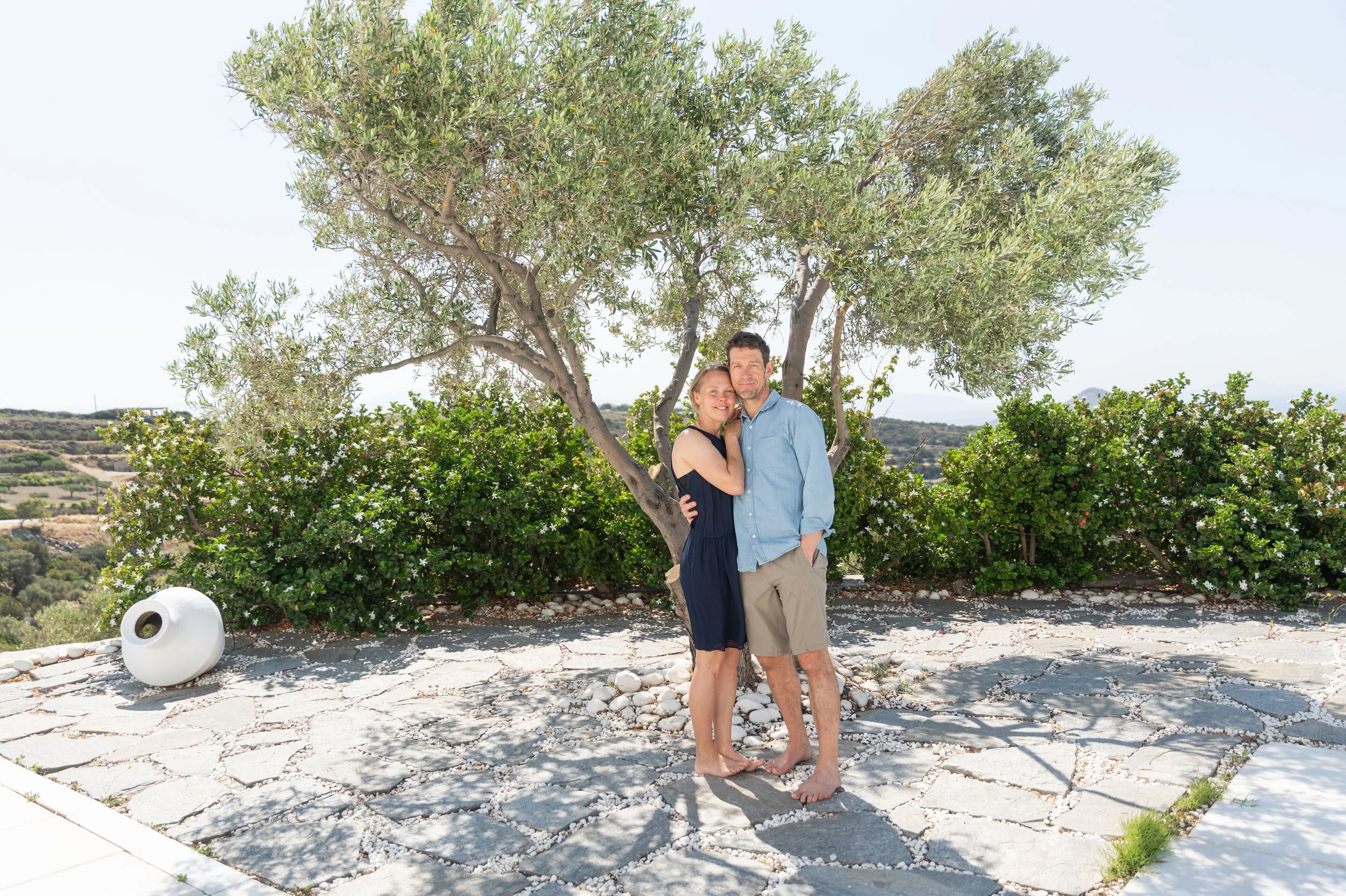 A couple standing close together in front of a leafy tree on a sunny patio, with bushes and scenic landscape in the background. The woman wears a dark dress, and the man wears a light blue shirt and khaki shorts.