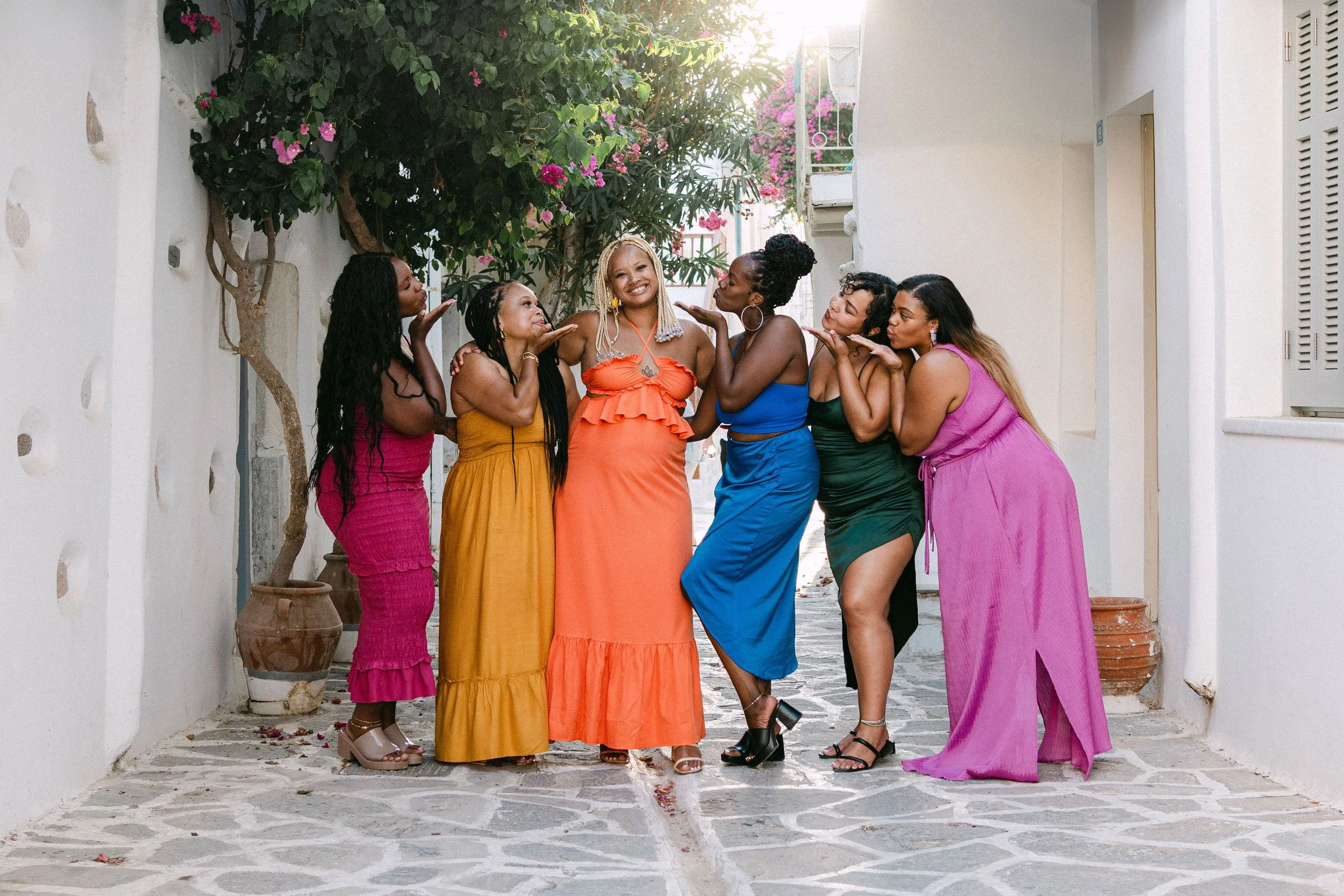 Group of six women in colorful dresses standing on cobblestone street, smiling and posing affectionately with each other.