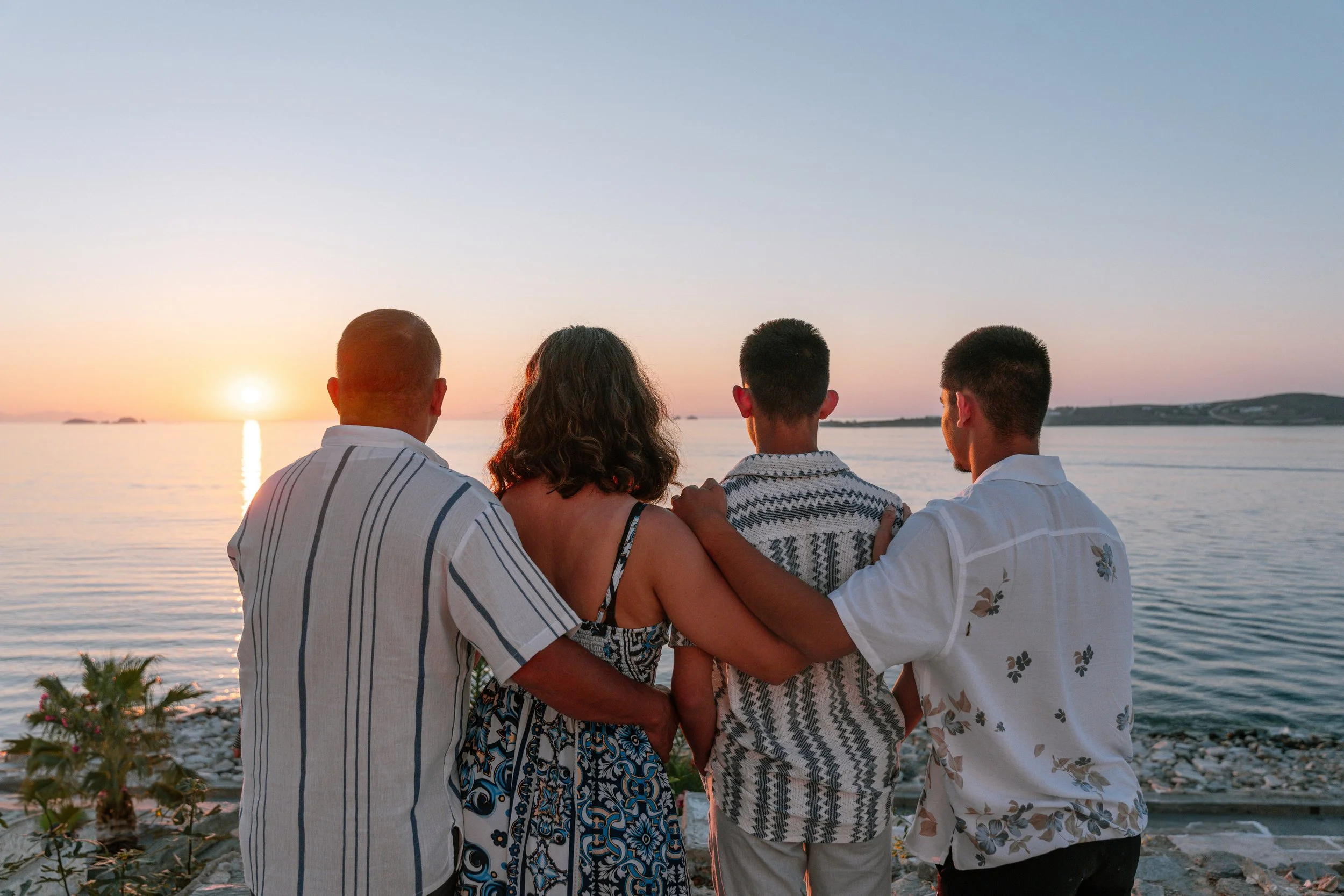 Four people, two men, one woman, and a young boy, sit on rocks by the water, watching the sunset over the ocean on a clear evening.