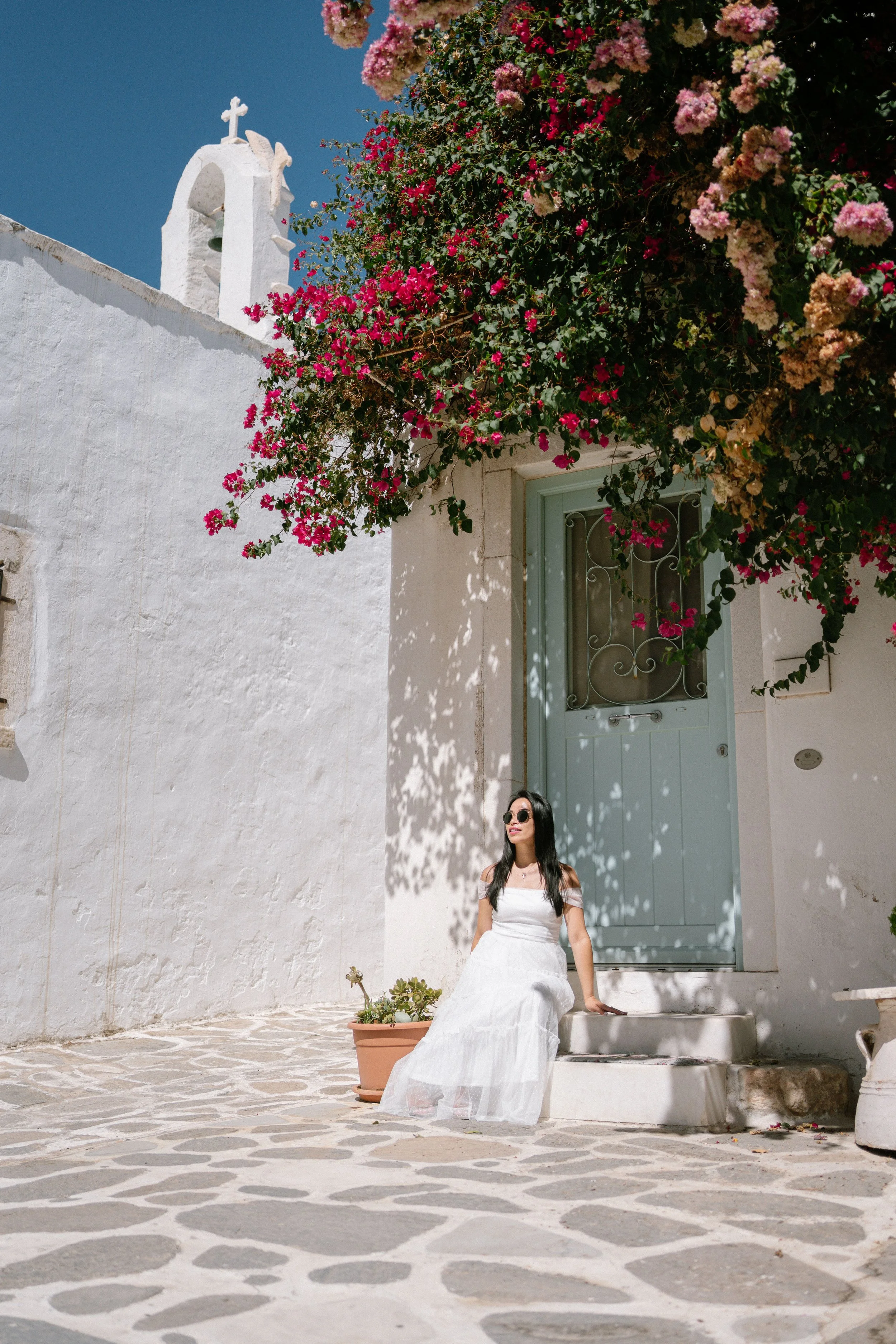 A woman in a white dress and sunglasses sitting on the steps in front of a white building with a light blue door, a large flowering bougainvillea tree overhead, and a small pot with plants nearby, under a clear blue sky.