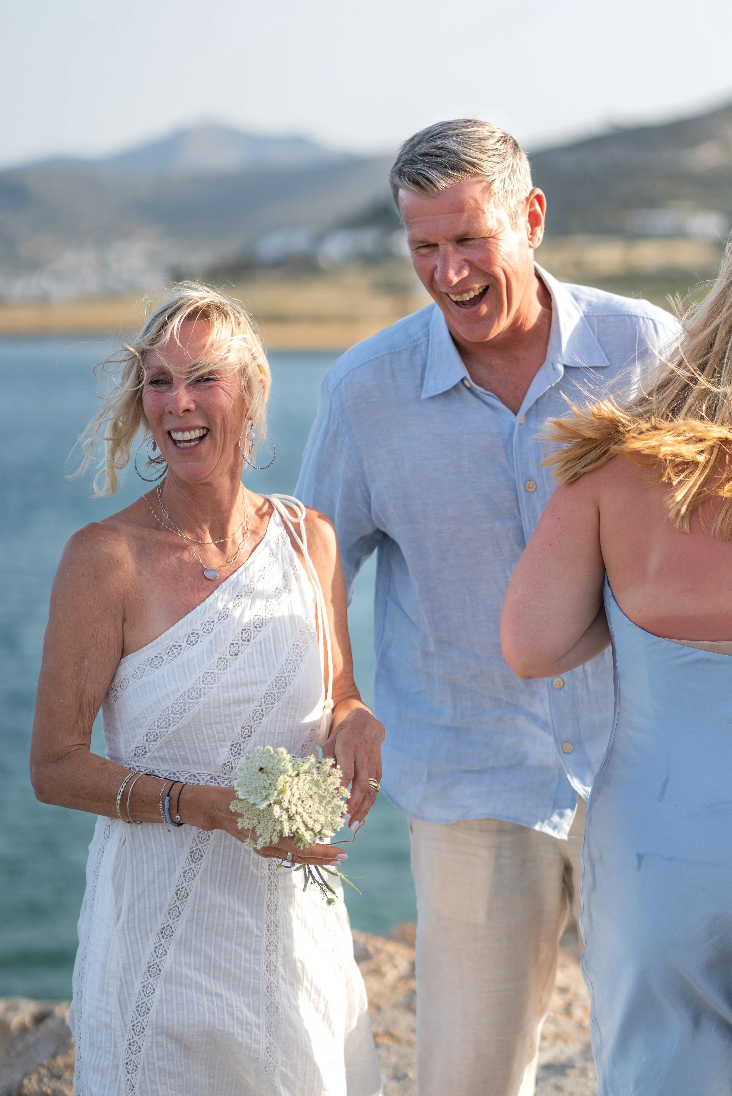 A group of people, including a woman in a white dress holding a bouquet and a man in a light blue shirt, smiling and standing near a body of water, with mountains in the background.