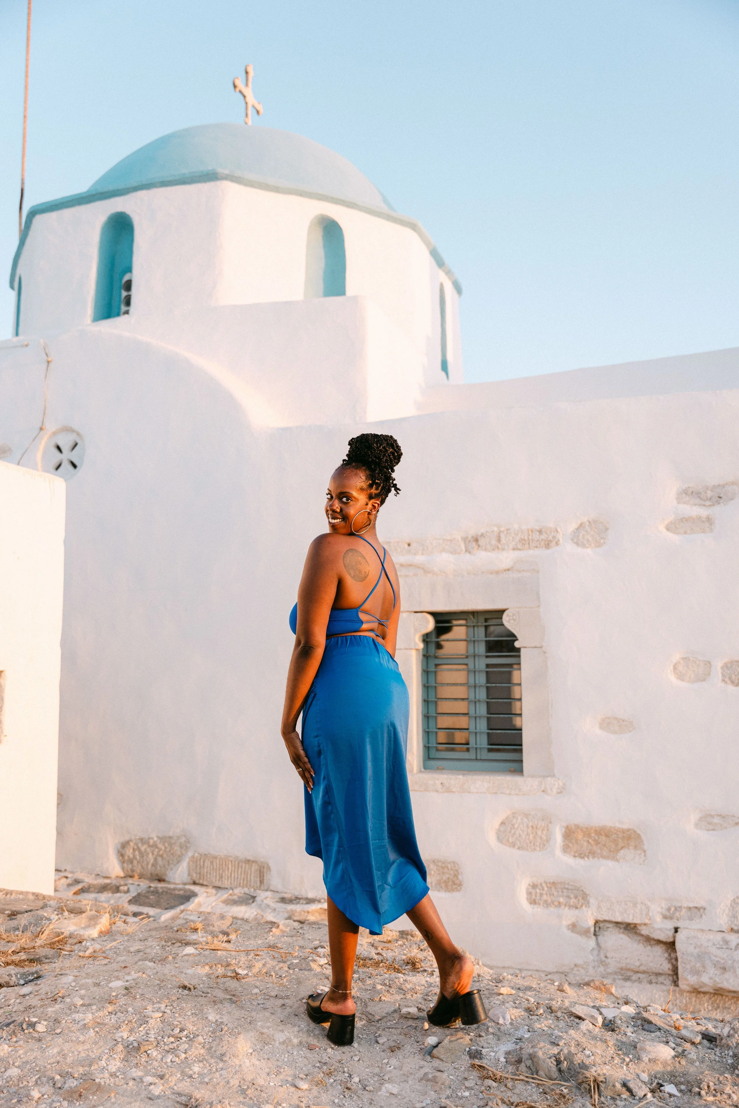 A woman smiling in a blue dress and black heeled sandals standing outdoors on a rocky path with white buildings and a church with a cross on top in the background.