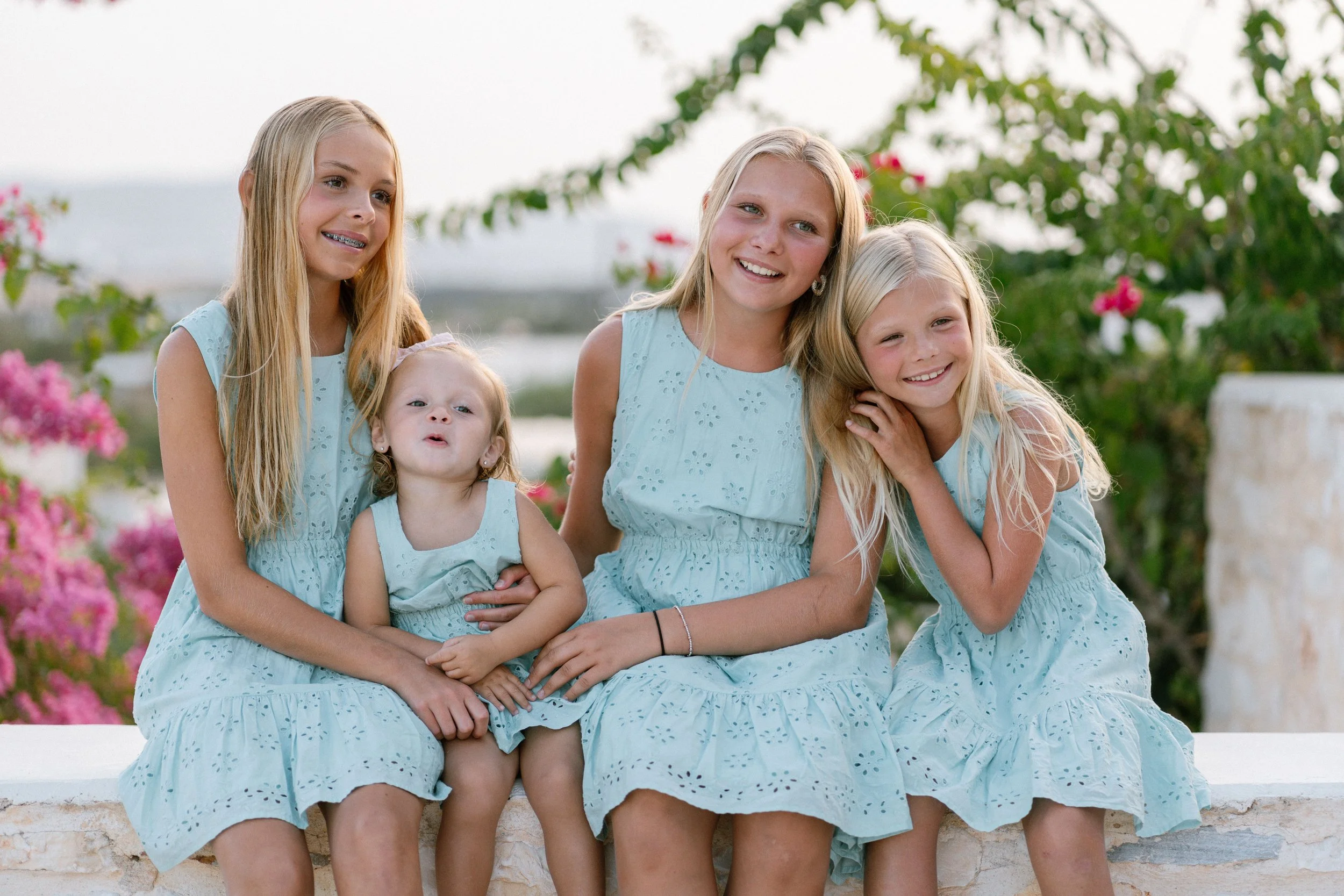 Five young girls in matching light blue dresses sitting outdoors on a stone wall, smiling and enjoying a sunny day with pink flowering bushes in the background.