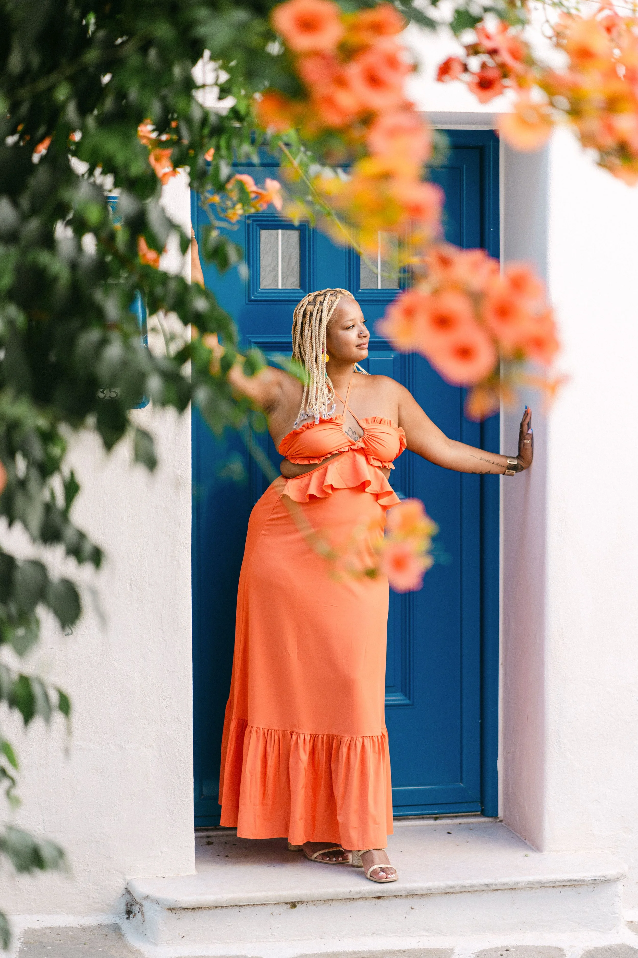 A woman in an orange dress with ruffles, standing at a blue door, framed by pink flowers and green leaves.