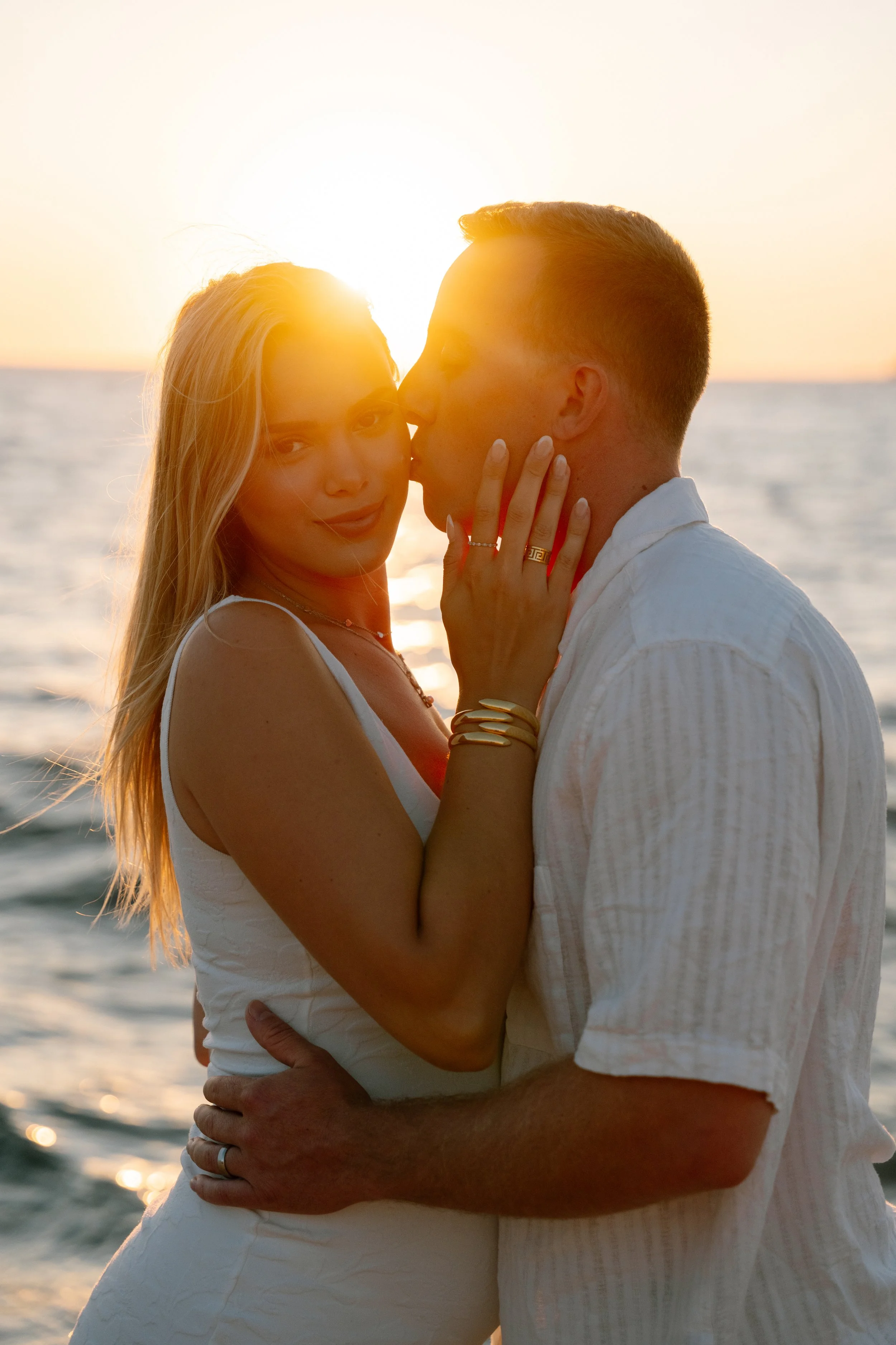 A couple is embracing and kissing at the beach during sunset, with the sun setting over the ocean in the background.