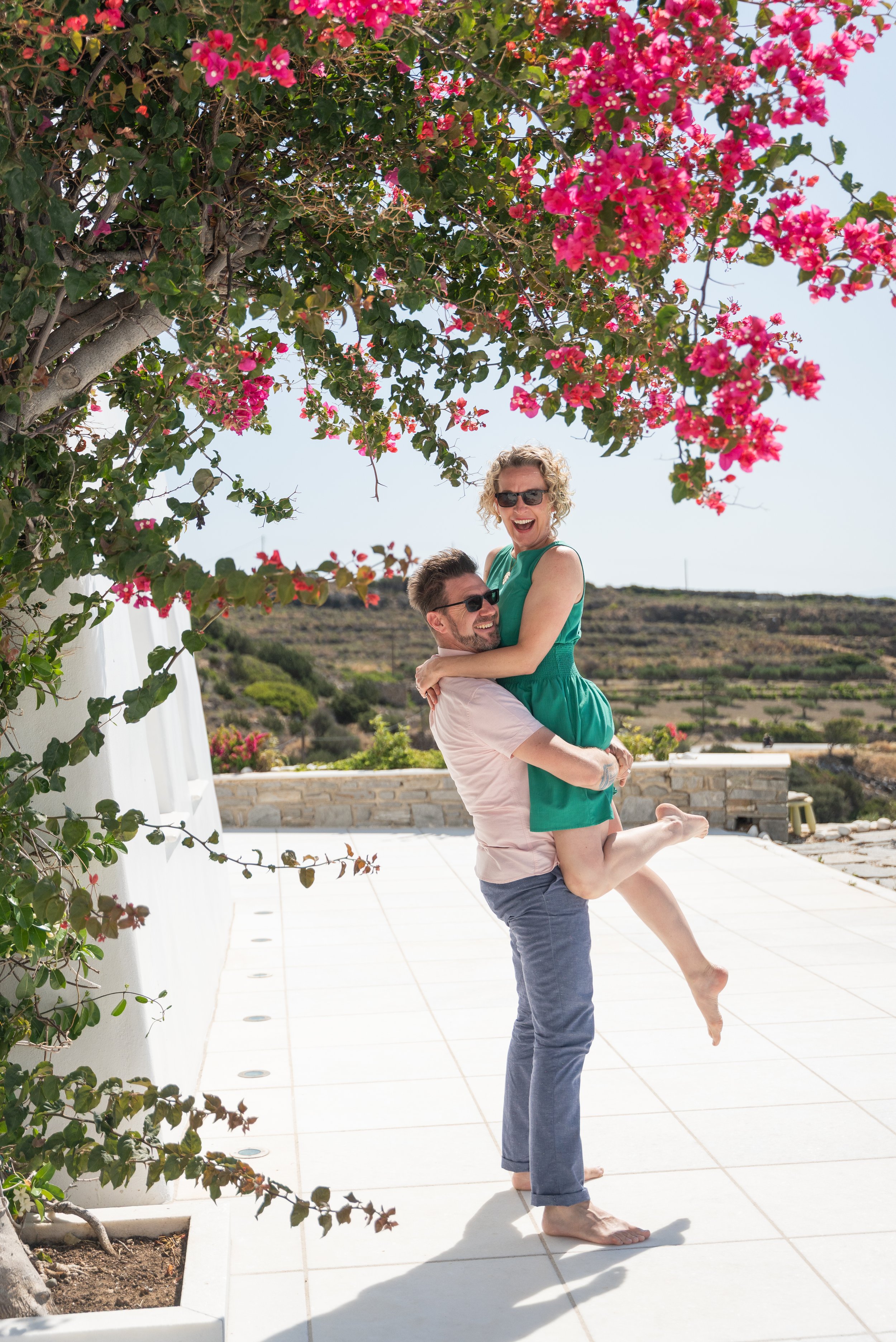 A man holding a woman in his arms, both smiling and wearing sunglasses, on a sunny terrace with pink flowering bushes and open landscape in the background.