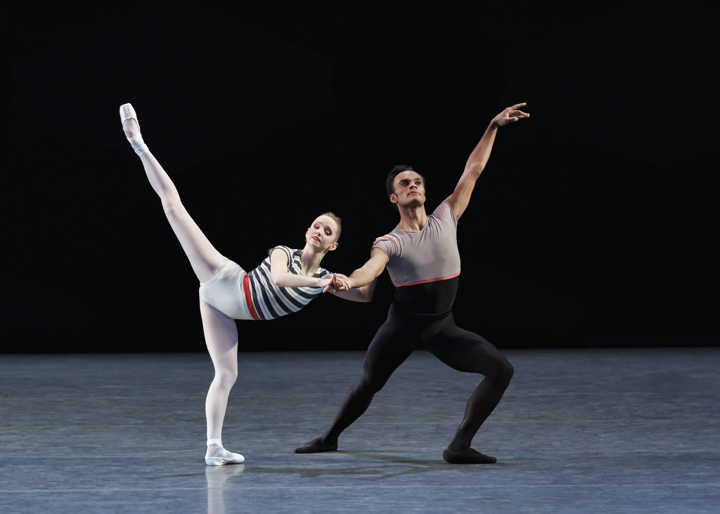 Ballet dancers, a female and a male, perform on stage. The female dancer is holding the male dancer’s hand while his other arm is raised. The stage background is black.