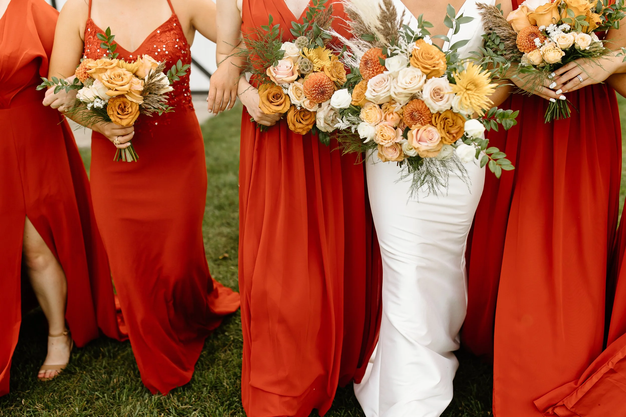 Close-up of bridesmaids holding bouquets of flowers, standing outdoors on grass, wearing red dresses, with one woman in a white dress at a Wisconsin barn wedding venue