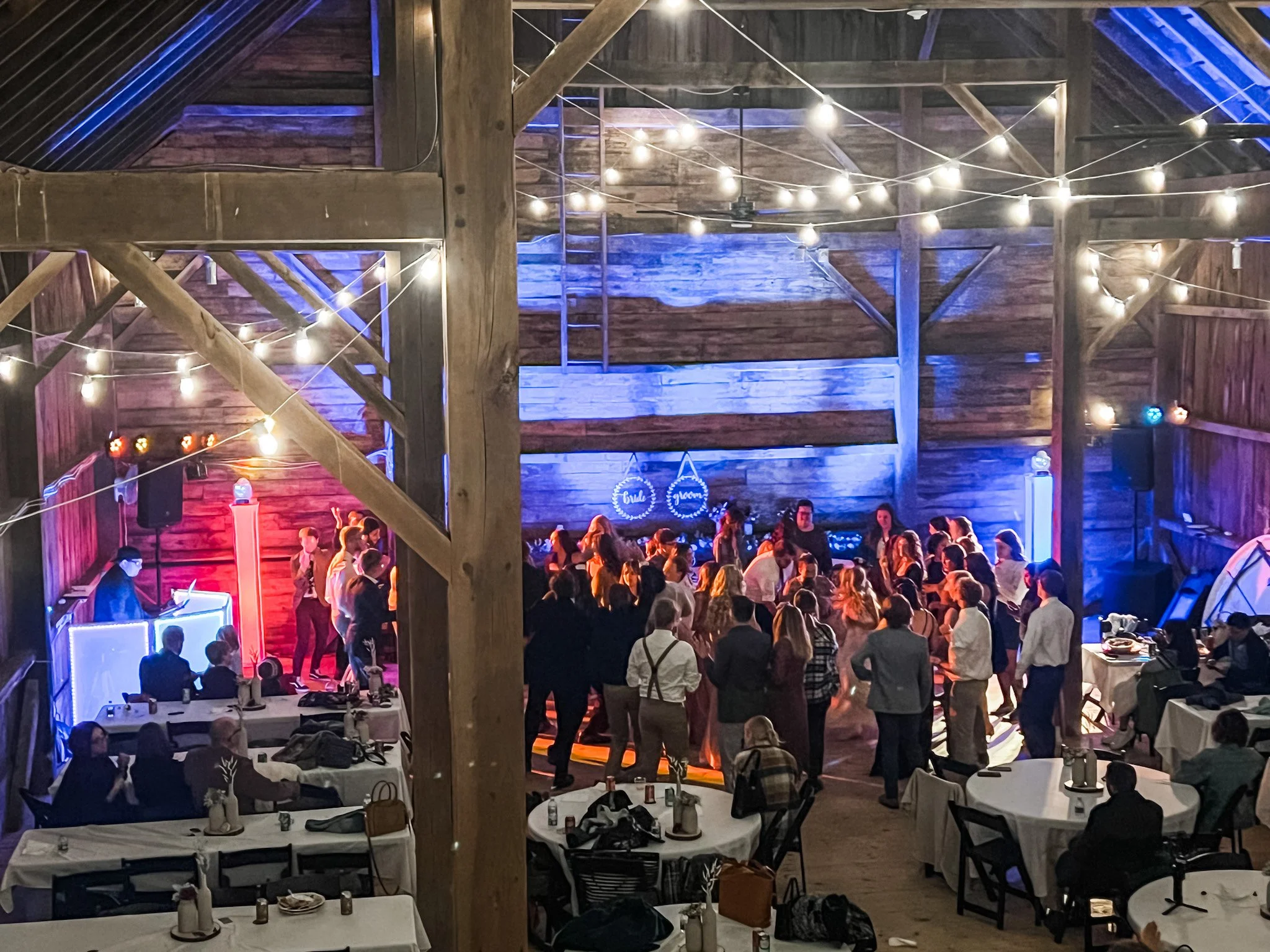 Inside a wooden barn decorated with string lights, a large group of people is gathered for a wedding or celebration, with tables and a dance floor area visible at this new richmond wedding venue