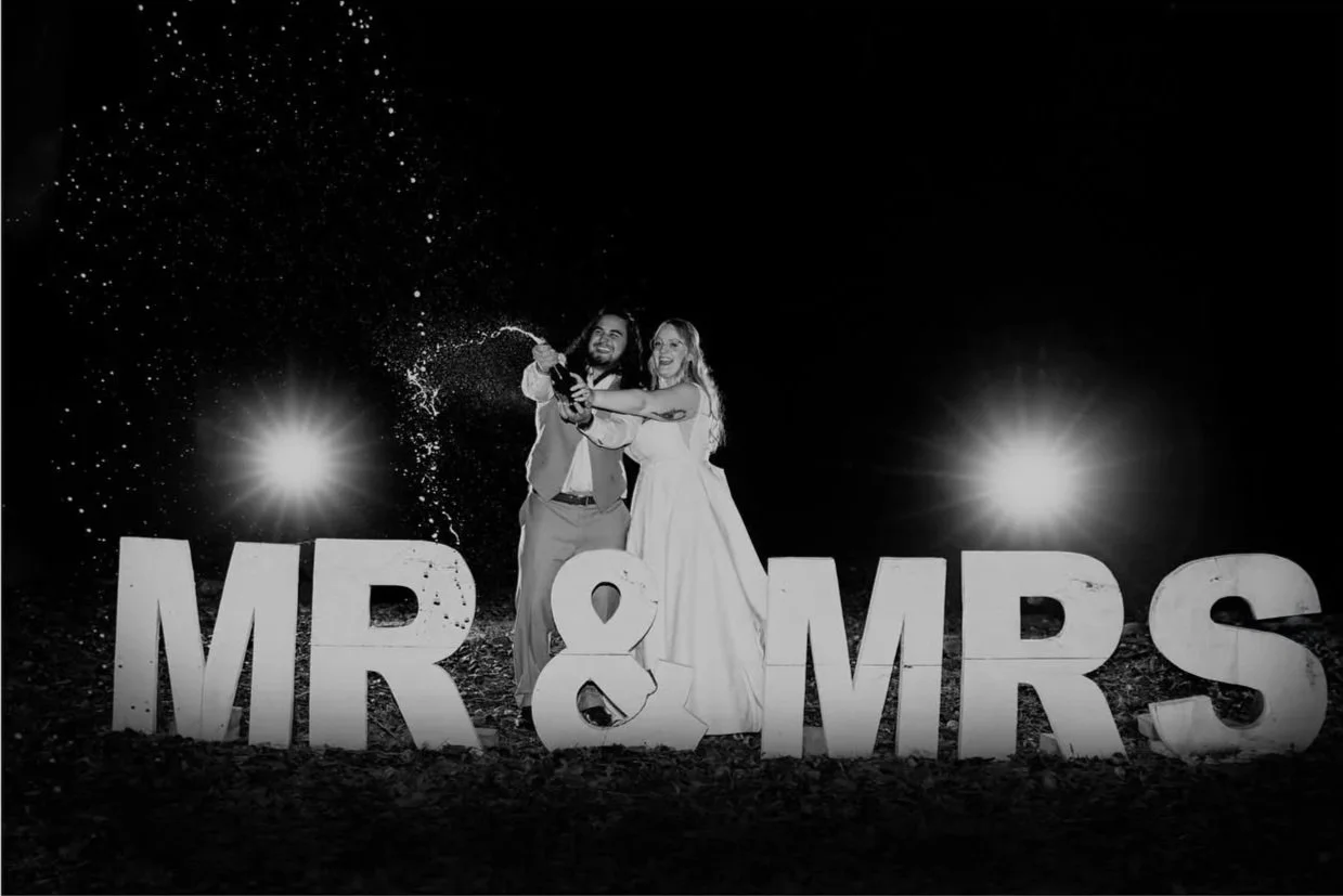 A bride and groom celebrating at night with large letters spelling 'MR & MRS' in the foreground. They are spraying champagne, and bright lights are illuminating the scene.
