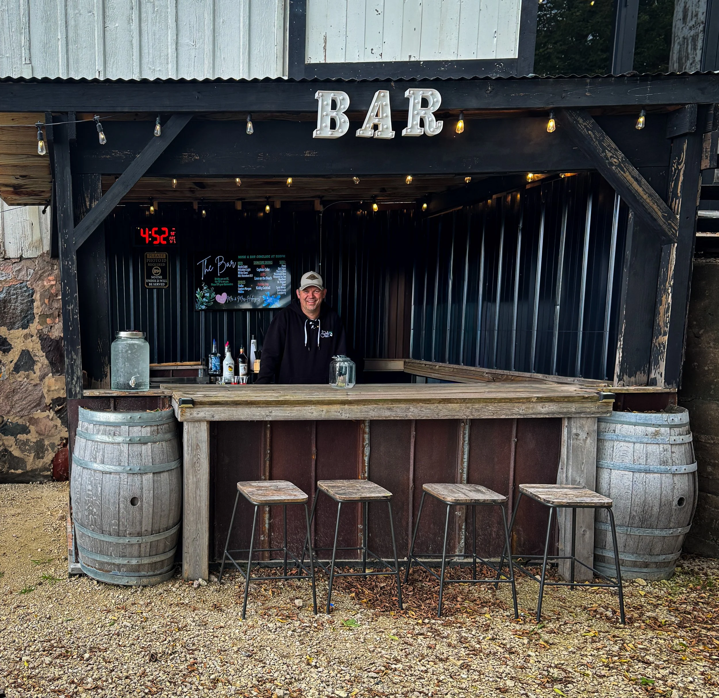 Outdoor bar with a man standing behind the counter, decorated with string lights and a sign that reads 'BAR' in large white letters. There are bottles and drink dispensers on the counter at this New Richmond wedding ven