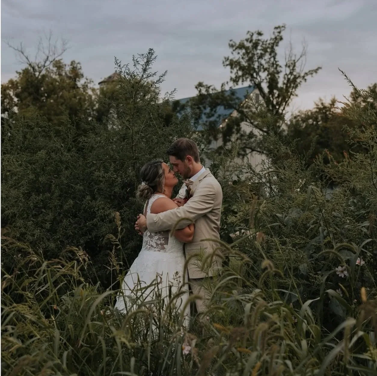 A couple dressed in wedding attire embrace each other among tall grass and bushes outdoors, with trees and a building in the background during the evening.