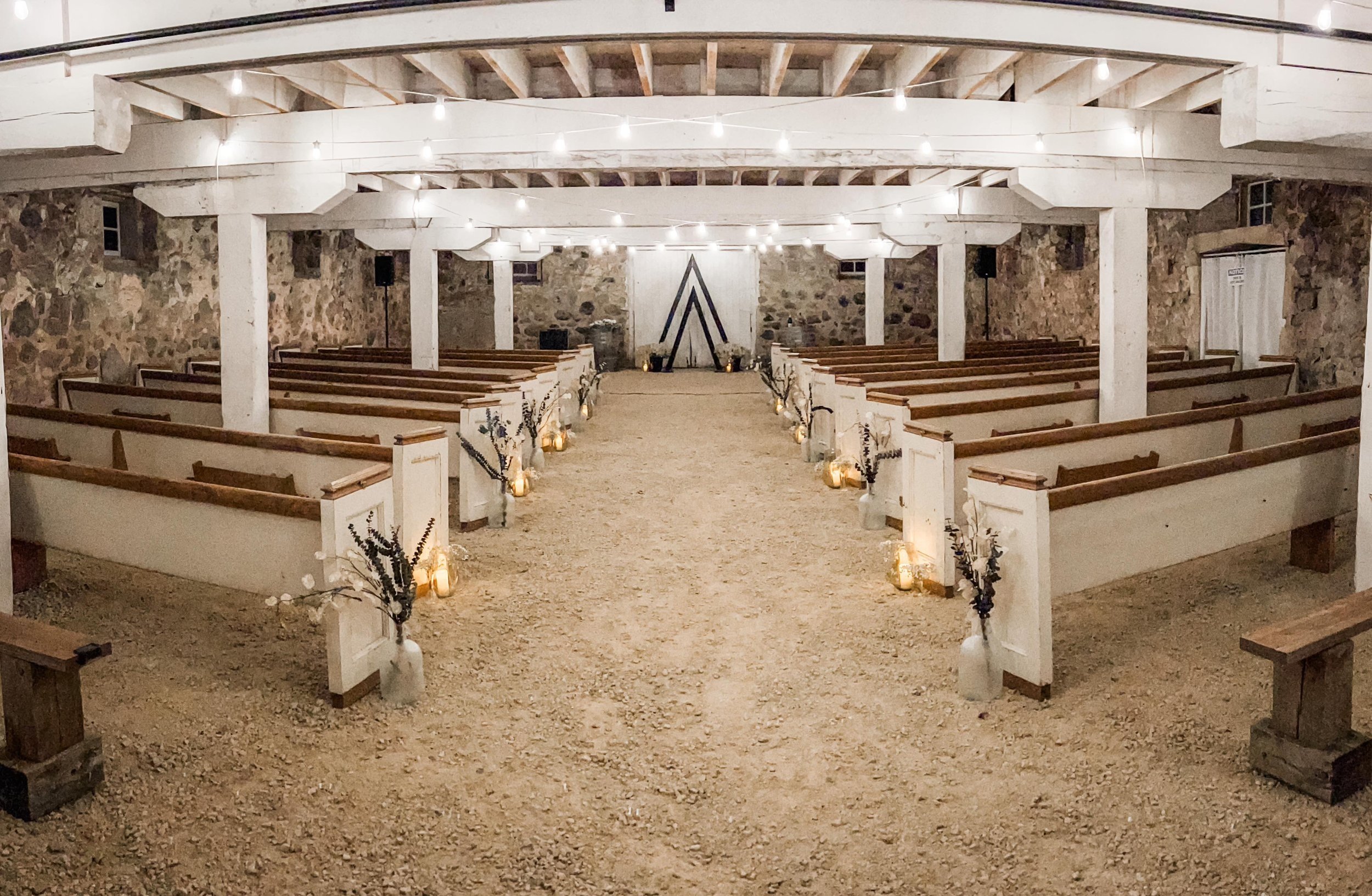A rustic indoor wedding ceremony space with wooden pews, a decorated aisle, string lights hanging from the ceiling, and a simple backdrop with a black geometric design on a white wooden panel.