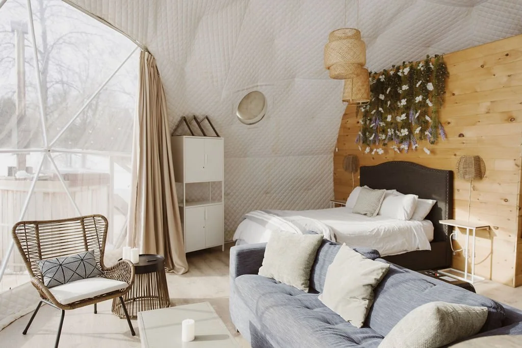 A cozy bedroom with a sloped ceiling, a large window with curtains, a bed with white bedding and a dark headboard, a gray sofa with pillows, a wicker chair with a pillow, a small round side table, a white cabinet, and decorative hanging lights and wall art.