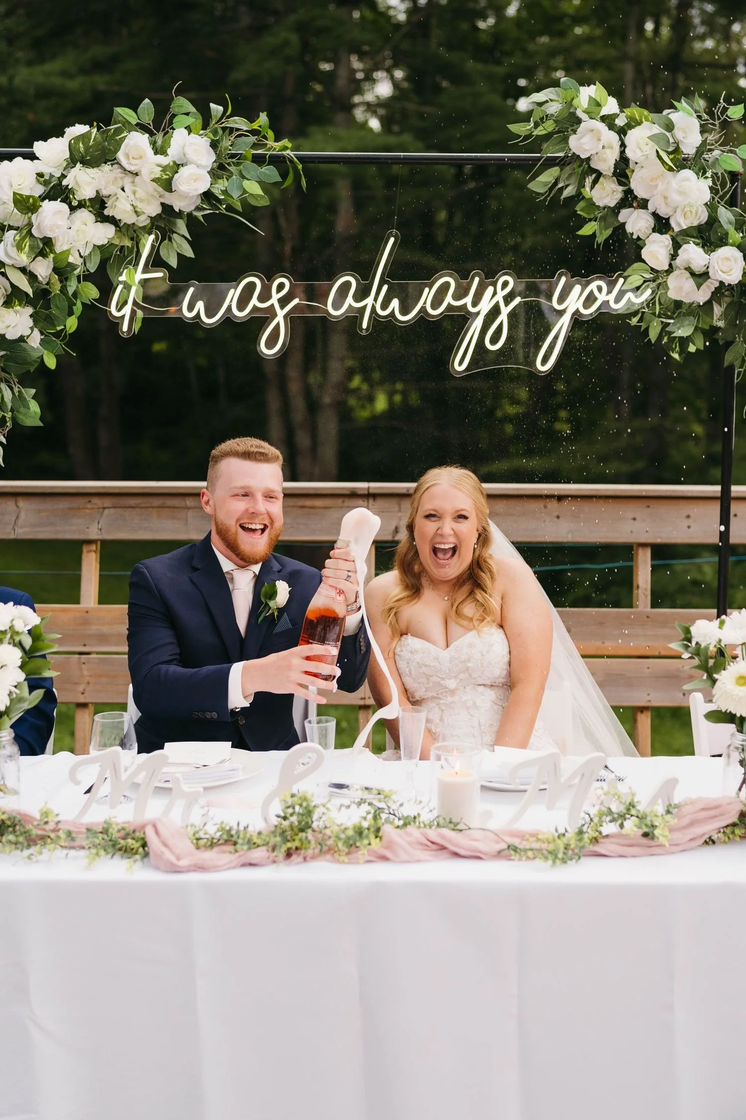 A bride and groom celebrating at their wedding reception, sitting at a decorated table outdoors. The groom is spraying champagne, and both are smiling and laughing. Behind them, a sign says 'It was always you' with floral decorations.