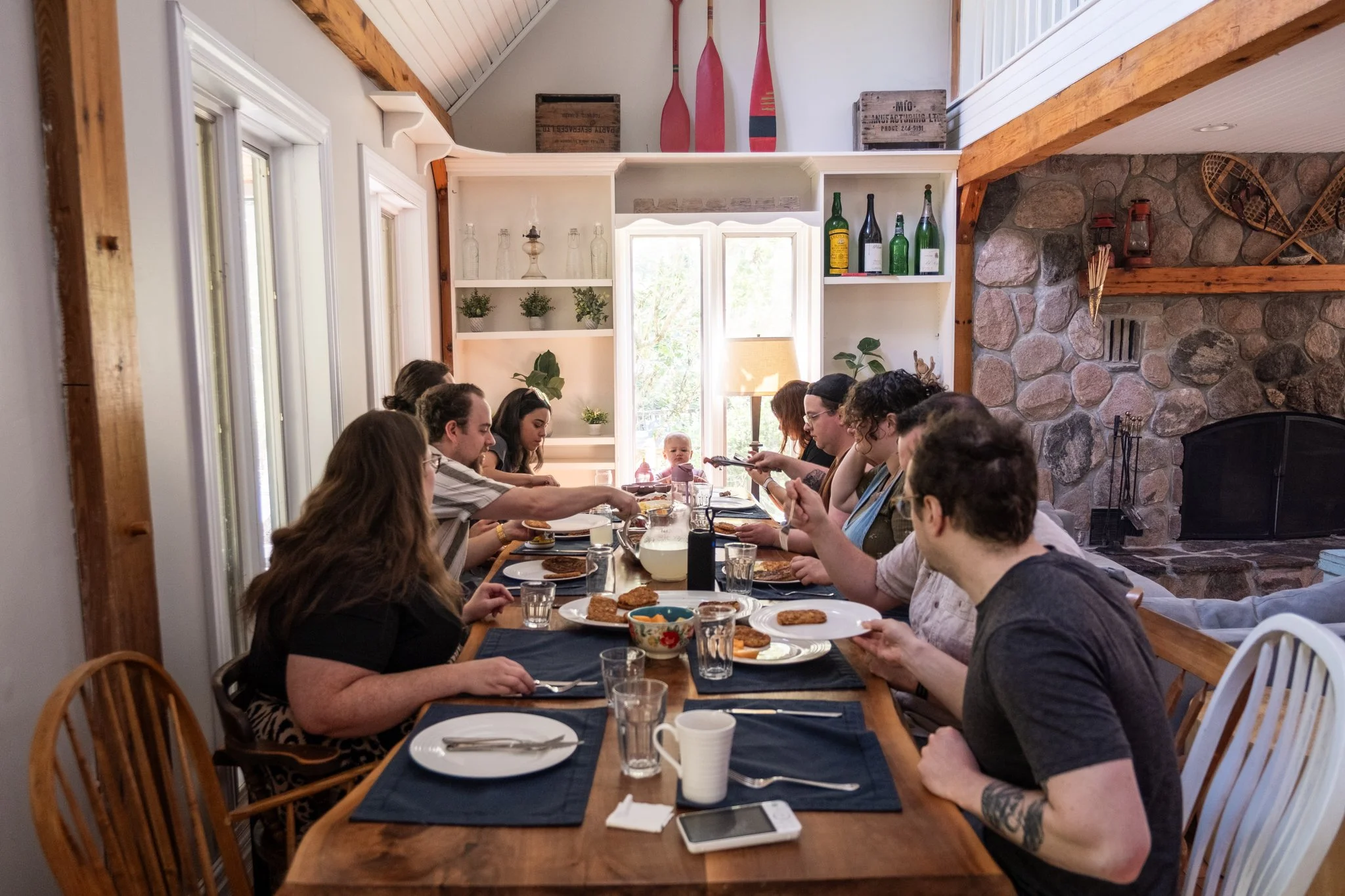 Multiple people sitting at a long dining table in a cozy, rustic room with a stone fireplace, sharing a meal together.