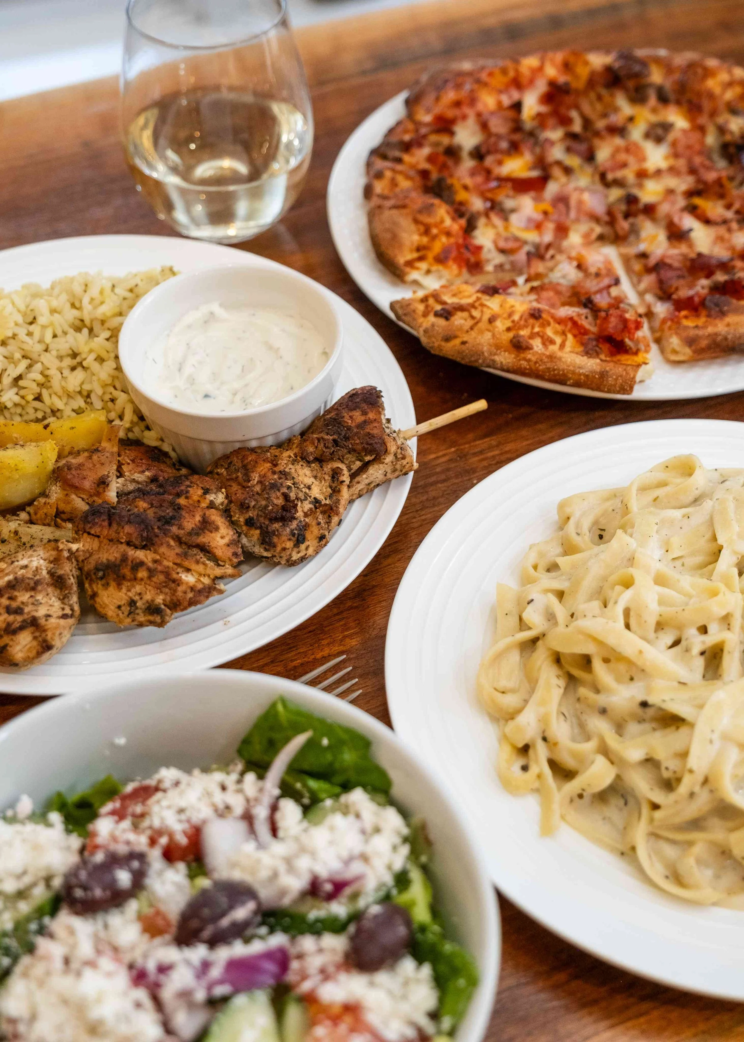 A meal with a slice of pizza, a bowl of pasta, grilled chicken skewers with a dipping sauce, rice, and a salad with feta cheese and olives, along with a glass of white wine on a wooden table.