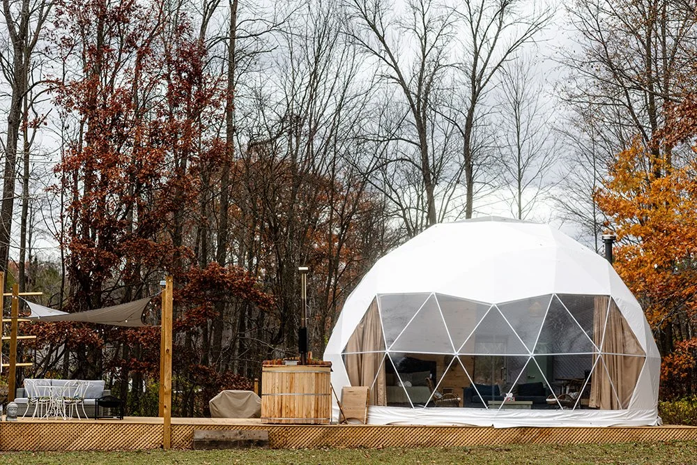 A backyard with a geodesic dome tent, outdoor seating area, and a hot tub, surrounded by trees with autumn foliage.