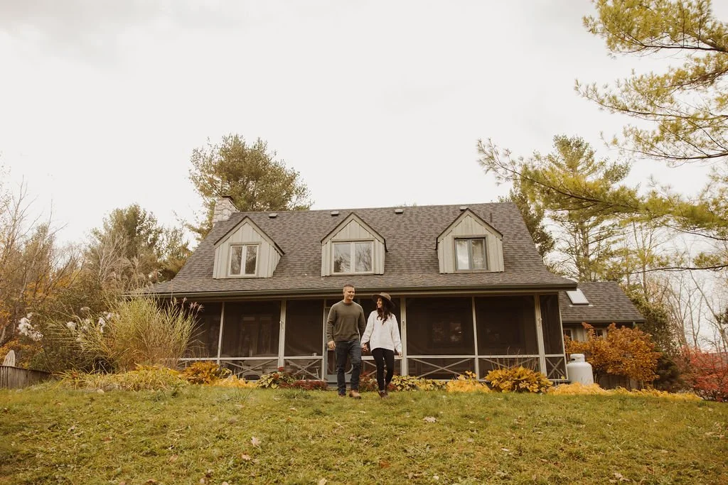 A couple walking in front of a large, two-story house with a screened porch and dormer windows, surrounded by trees with fall foliage.
