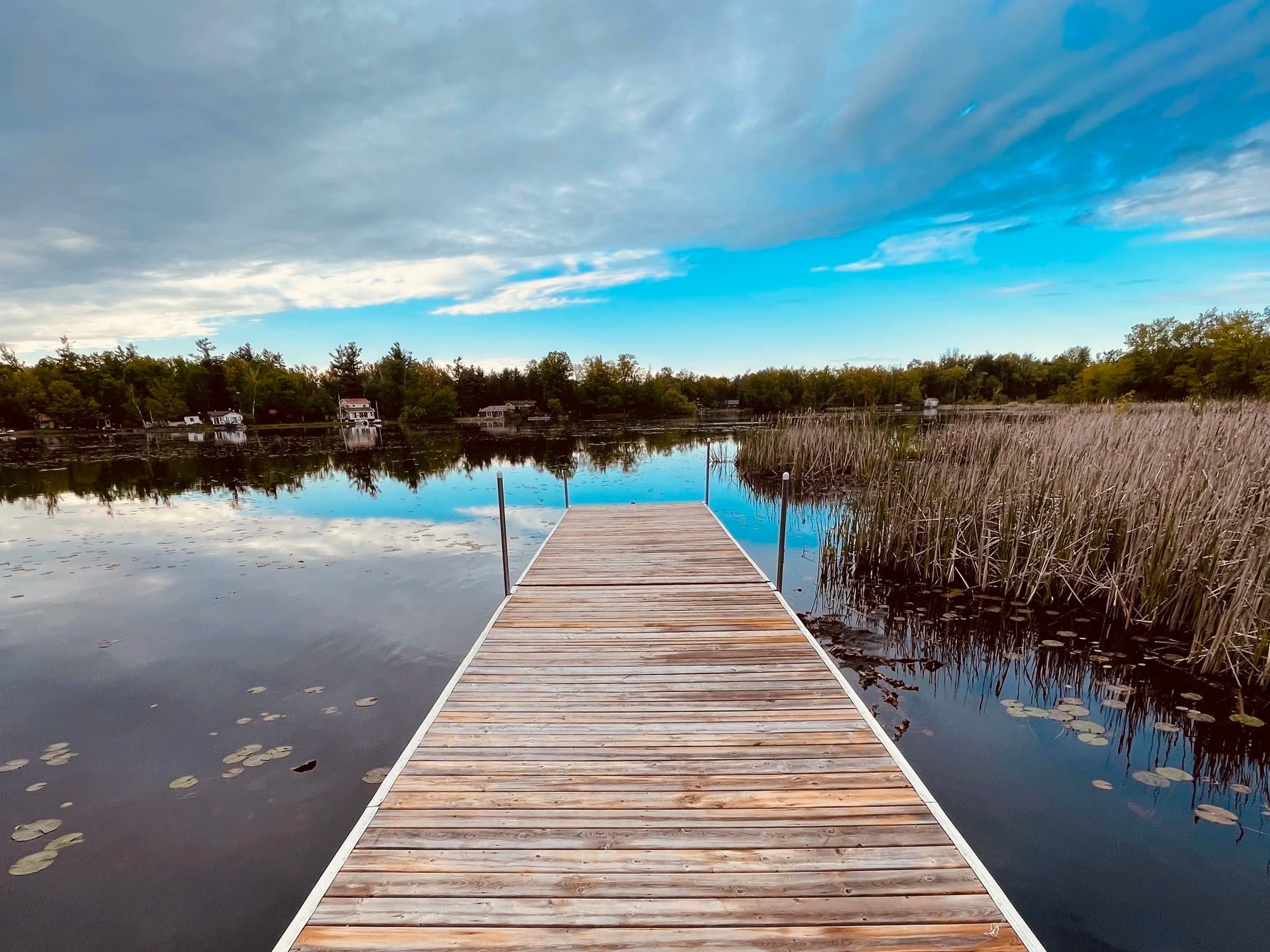 Scenic view of a wooden dock extending into a calm lake surrounded by trees in autumn colors, with cottages in the background at Marmora Retreats