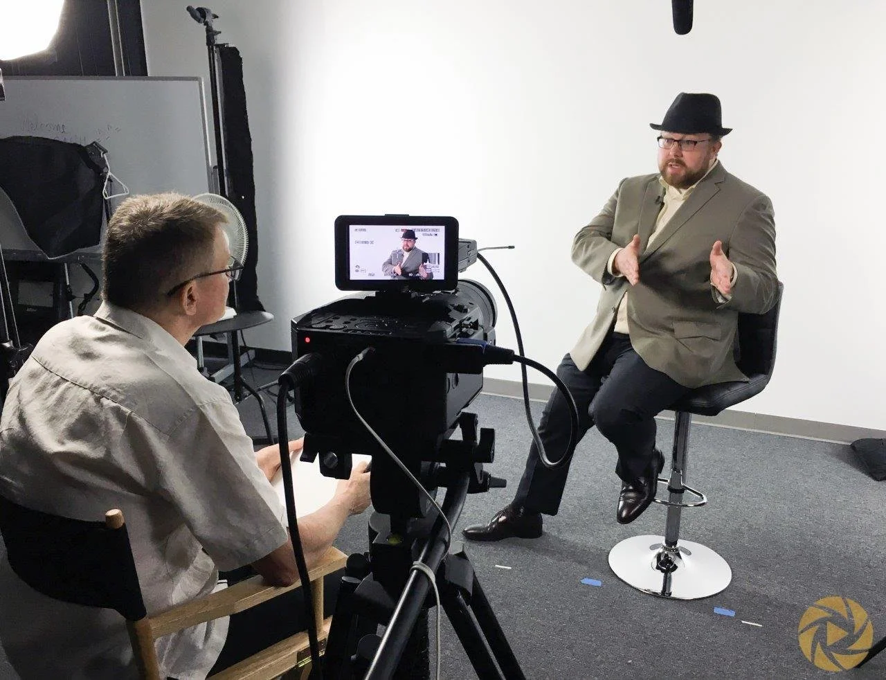 A man wearing a tan blazer, black fedora, glasses, and dark pants is sitting on a stool in a recording studio, gesturing with his hands while speaking. He is being filmed by a cameraman sitting in front of him, with a camera mounted on a tripod. The 