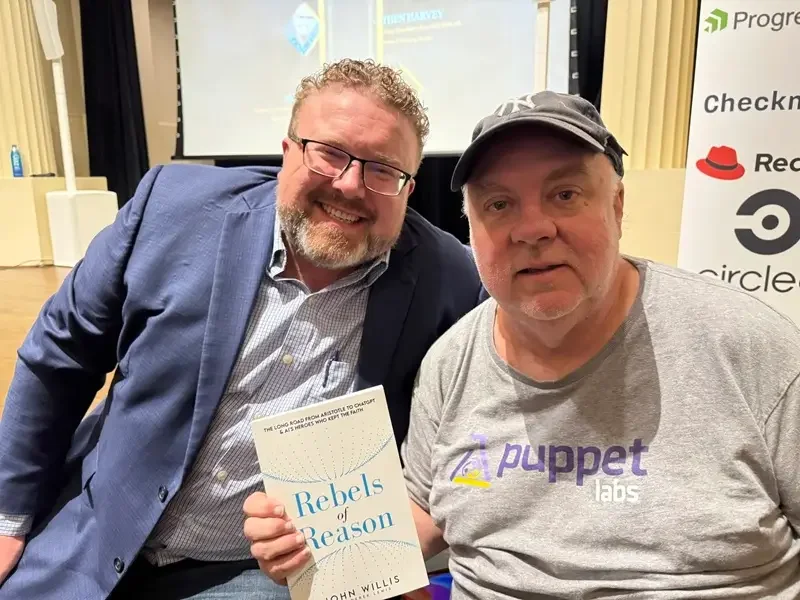 Business book ghostwriter Derek Lewis (left) in a blue blazer sitting on a presentation stage with client John Willis, who is holding a galley copy of his book Rebels of Reason.