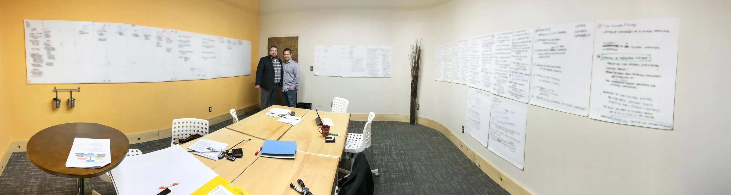 Two men standing in a meeting room with a long conference table, surrounded by white chairs, papers, notebooks, and office supplies. Whiteboards with writing on the walls, some of which are blurred, and a yellow accent wall with two hooks.