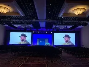 Large conference room with two big screens displaying a man with glasses, a hat, and a beard speaking with yellow subtitles. The stage has lighting equipment and a Microsoft logo in the center.