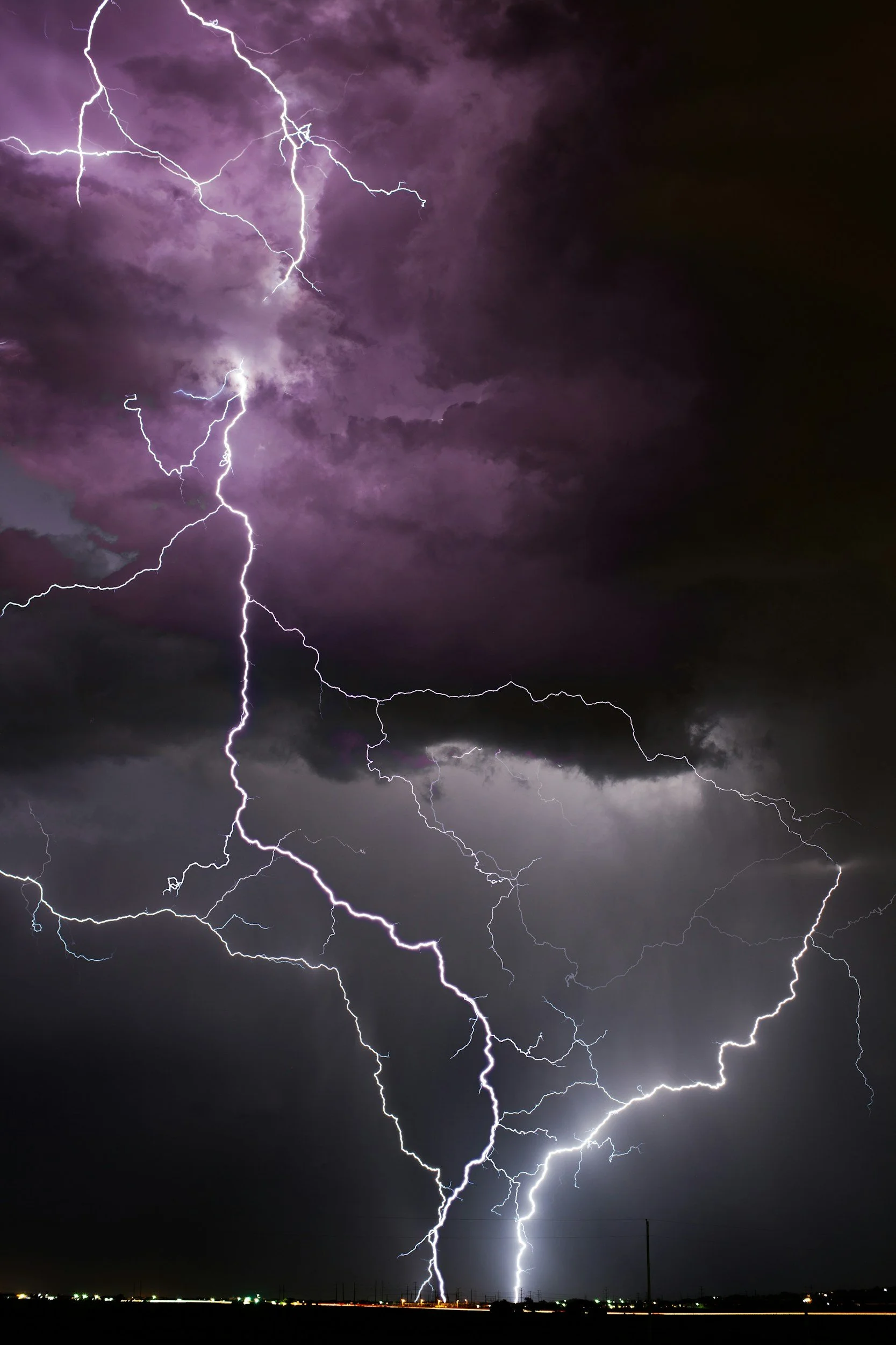 Multiple lightning bolts strike the ground during a thunderstorm at night, illuminating dark, purple-tinged clouds.