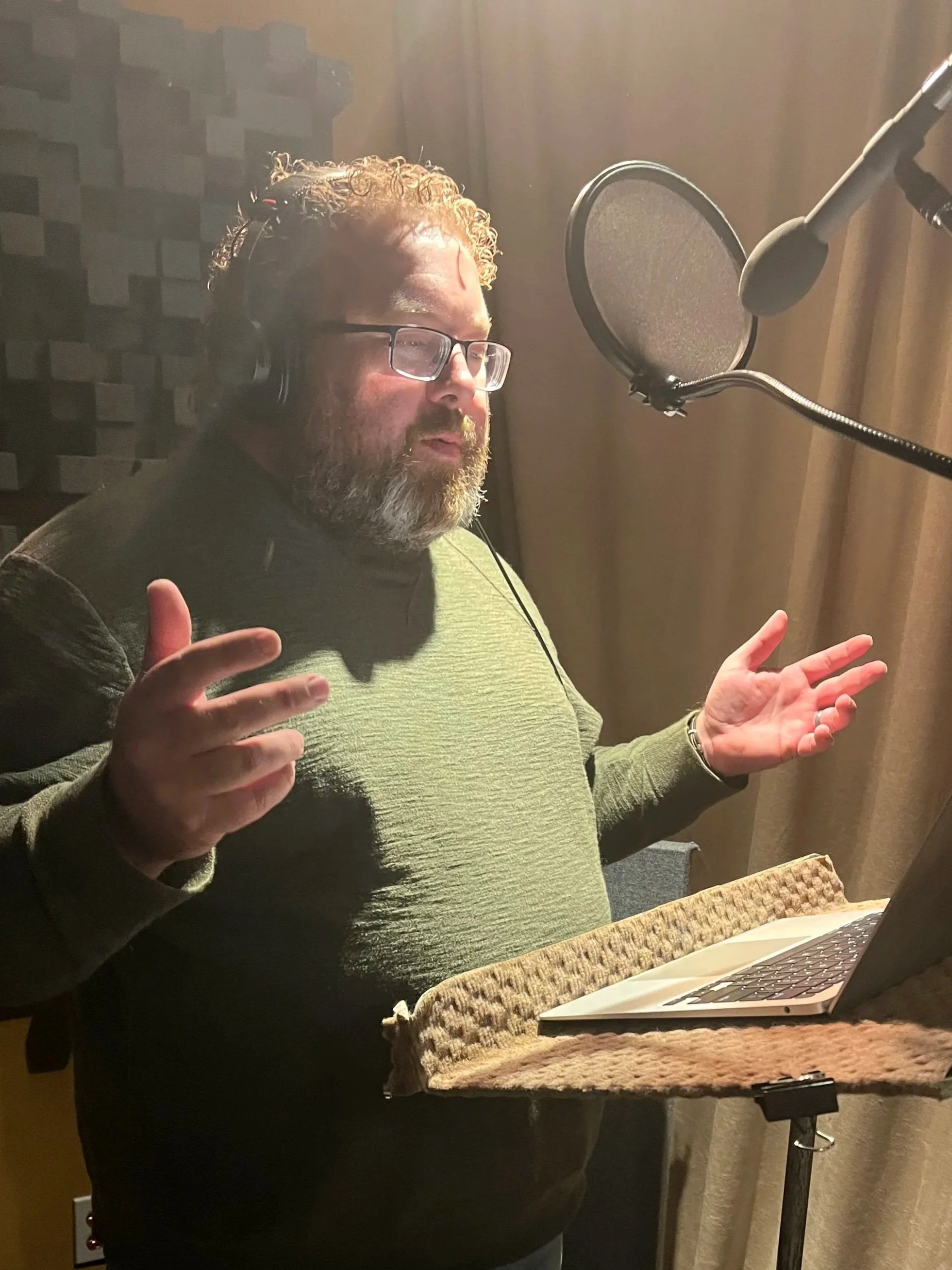 A man with glasses and a beard is singing or speaking into a microphone in a recording studio, with a laptop on a stand in front of him.