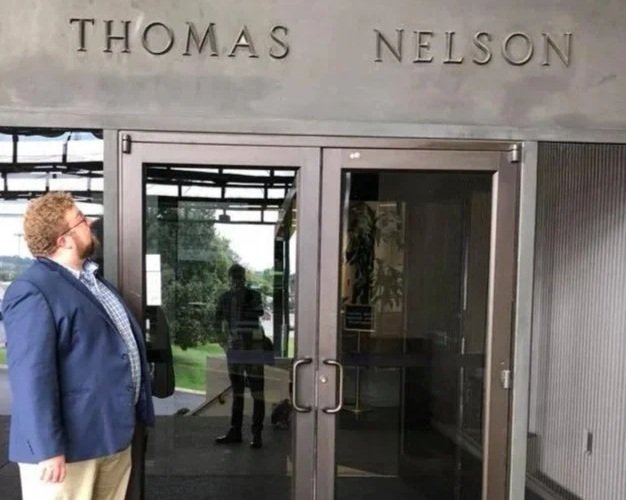 Man with curly hair and beard looking up at the Thomas Nelson building entrance with glass doors, reflecting trees, and a person inside.